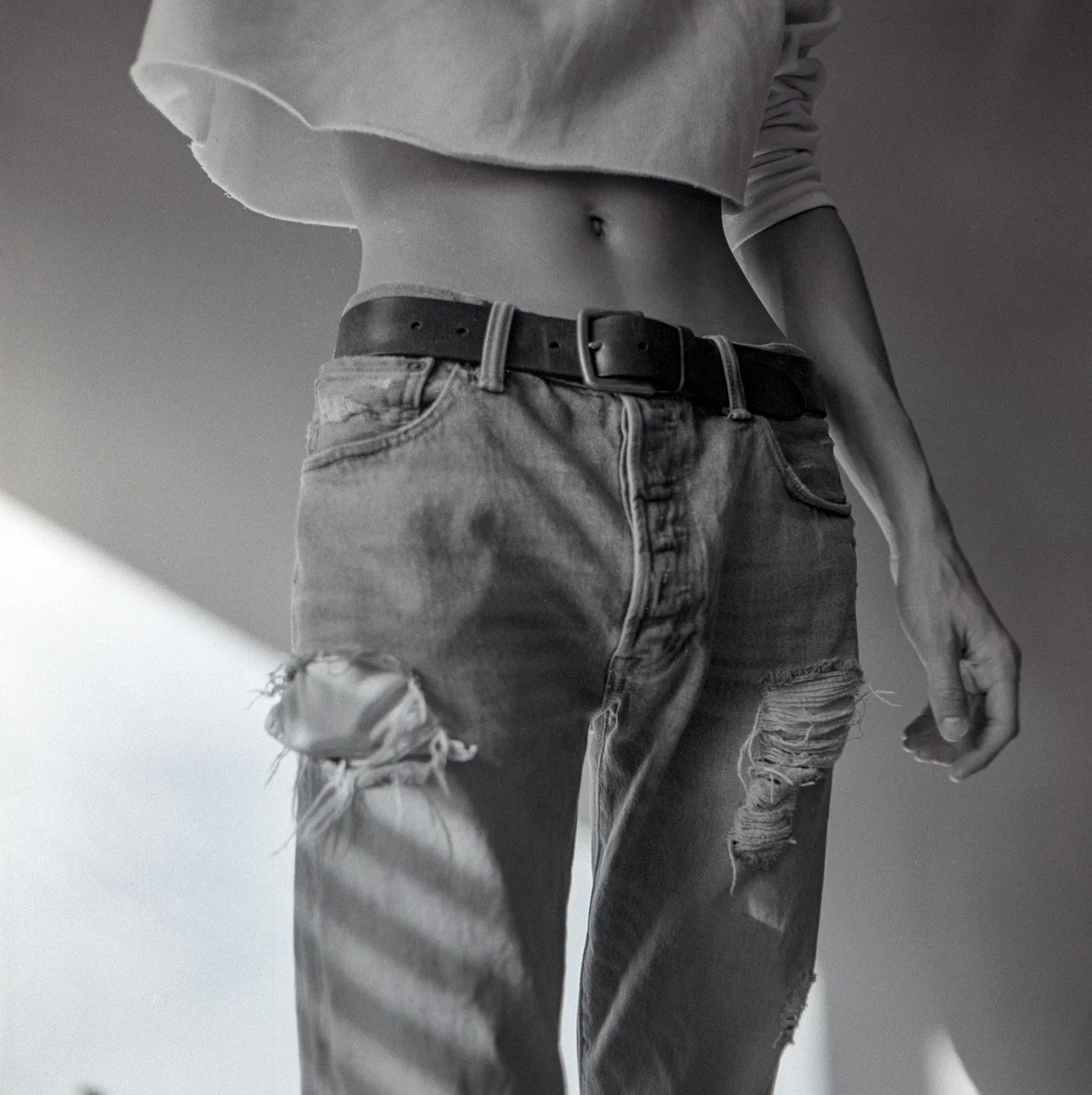 Black and white photo of a person wearing a cropped top, ripped jeans, and a belt, with a visible navel, standing in front of a plain background.