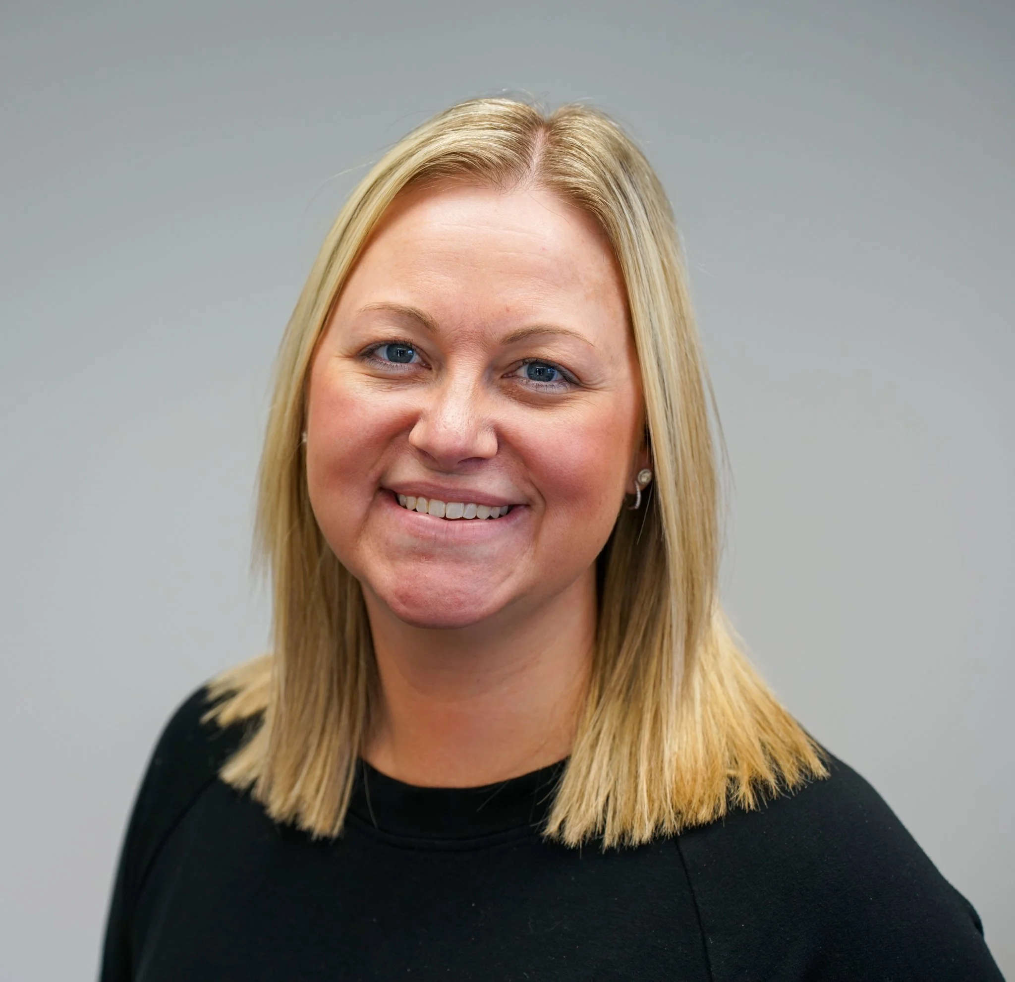 A woman with blonde hair smiling at the camera, wearing a black top, against a plain gray background.