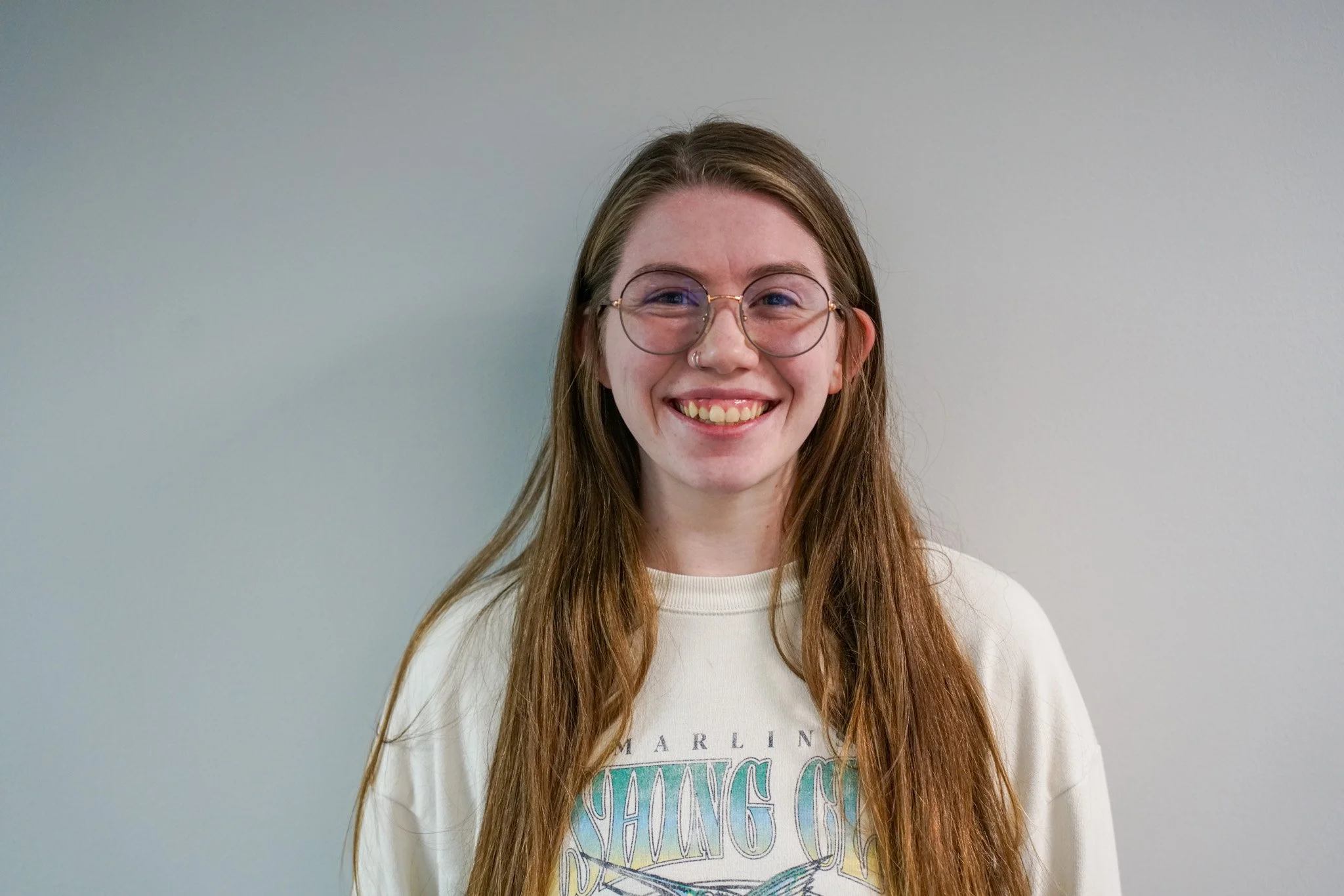 A smiling young woman with long red hair, wearing glasses and a light-colored sweatshirt, standing against a plain white wall.