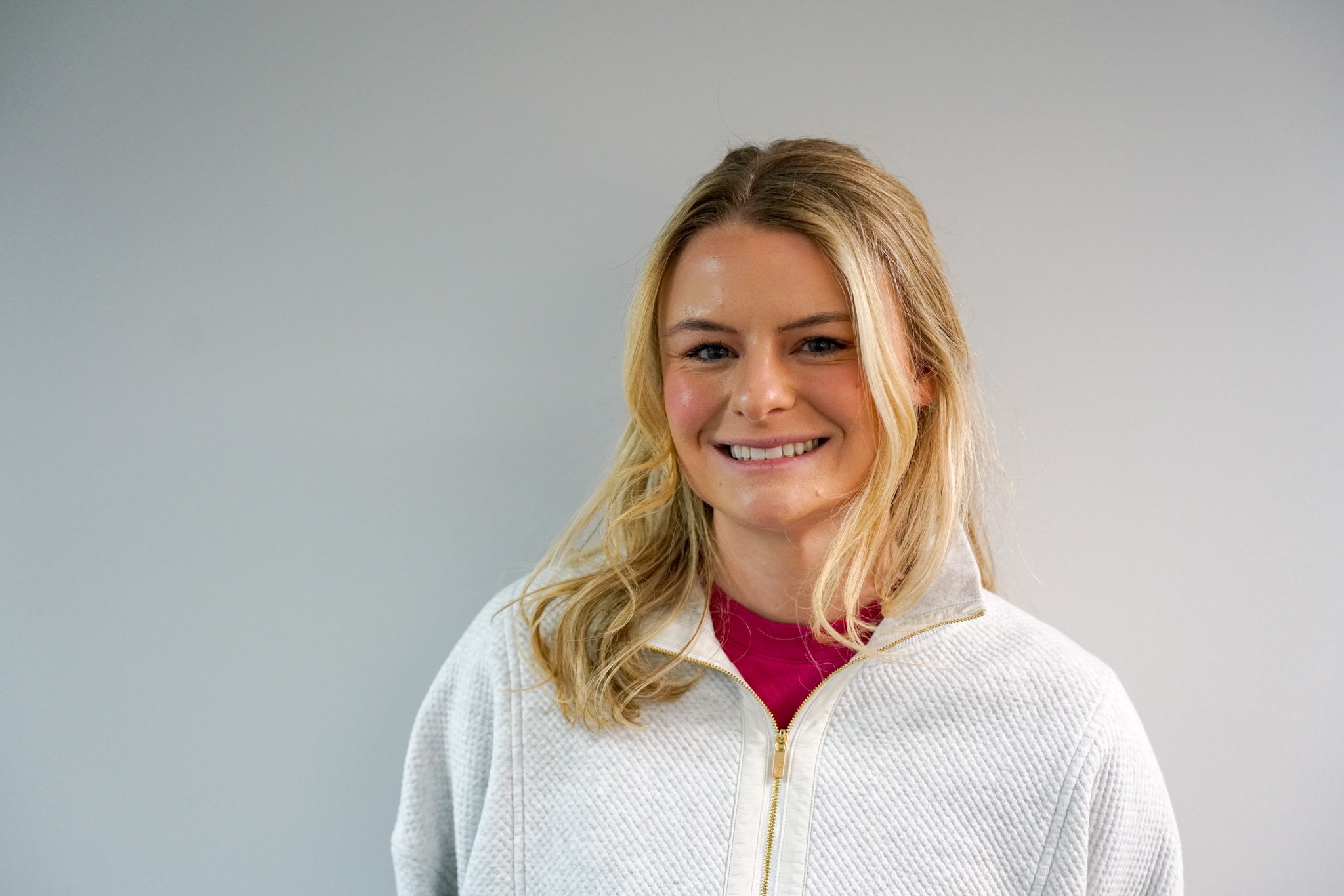 A woman with blonde hair smiling, wearing a white zip-up jacket with a pink shirt underneath, standing against a plain gray background.