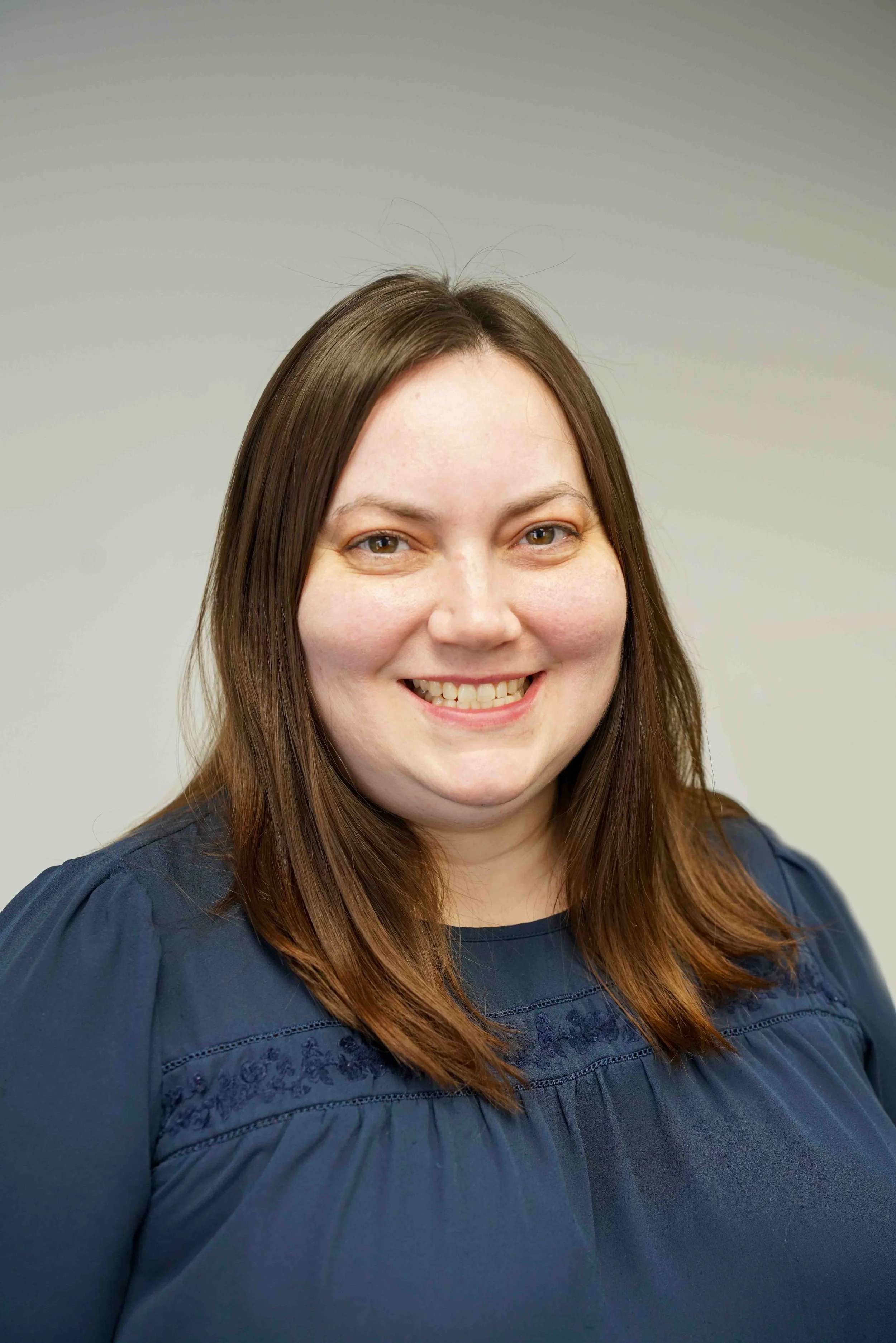 Portrait of a smiling woman with shoulder-length brown hair, wearing a dark blue blouse, against a plain light gray background.
