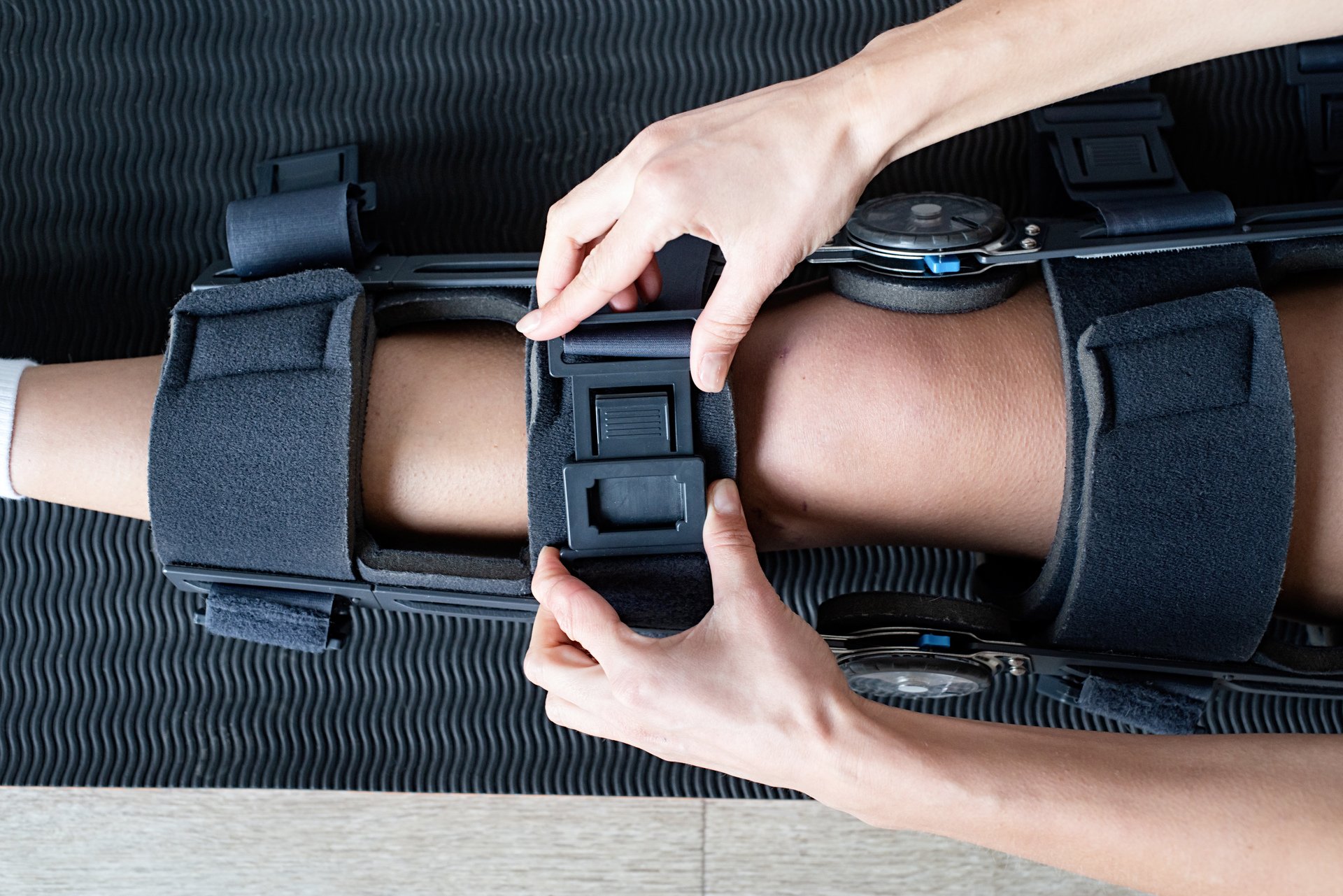 A person is securing a patient's leg with an orthopedic brace, with hands fastening straps around the leg on a black textured surface.