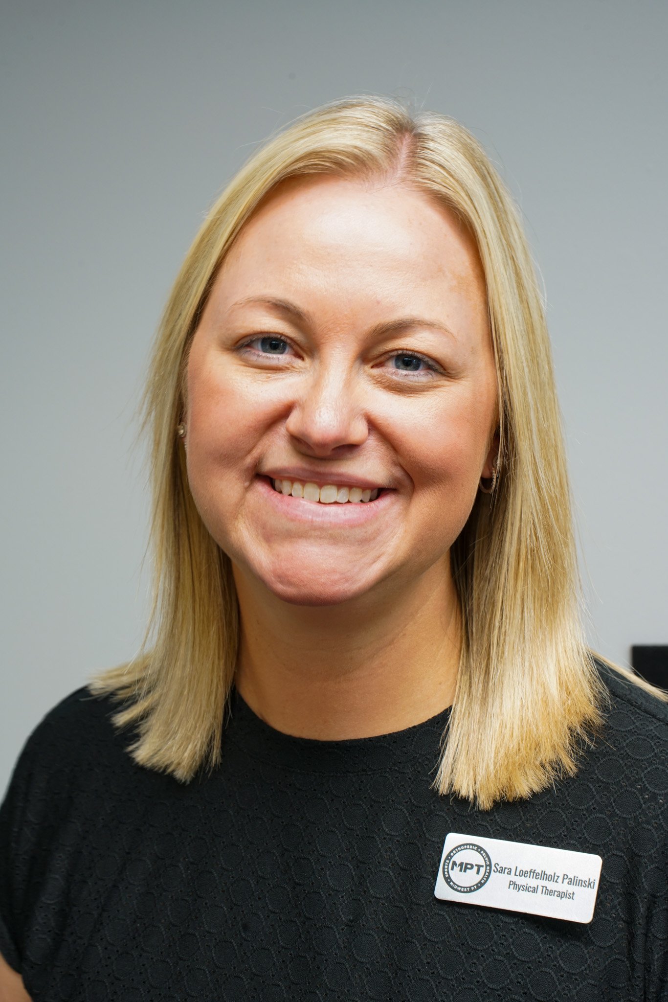 A woman with blonde hair smiling at the camera, wearing a black top, against a plain gray background.