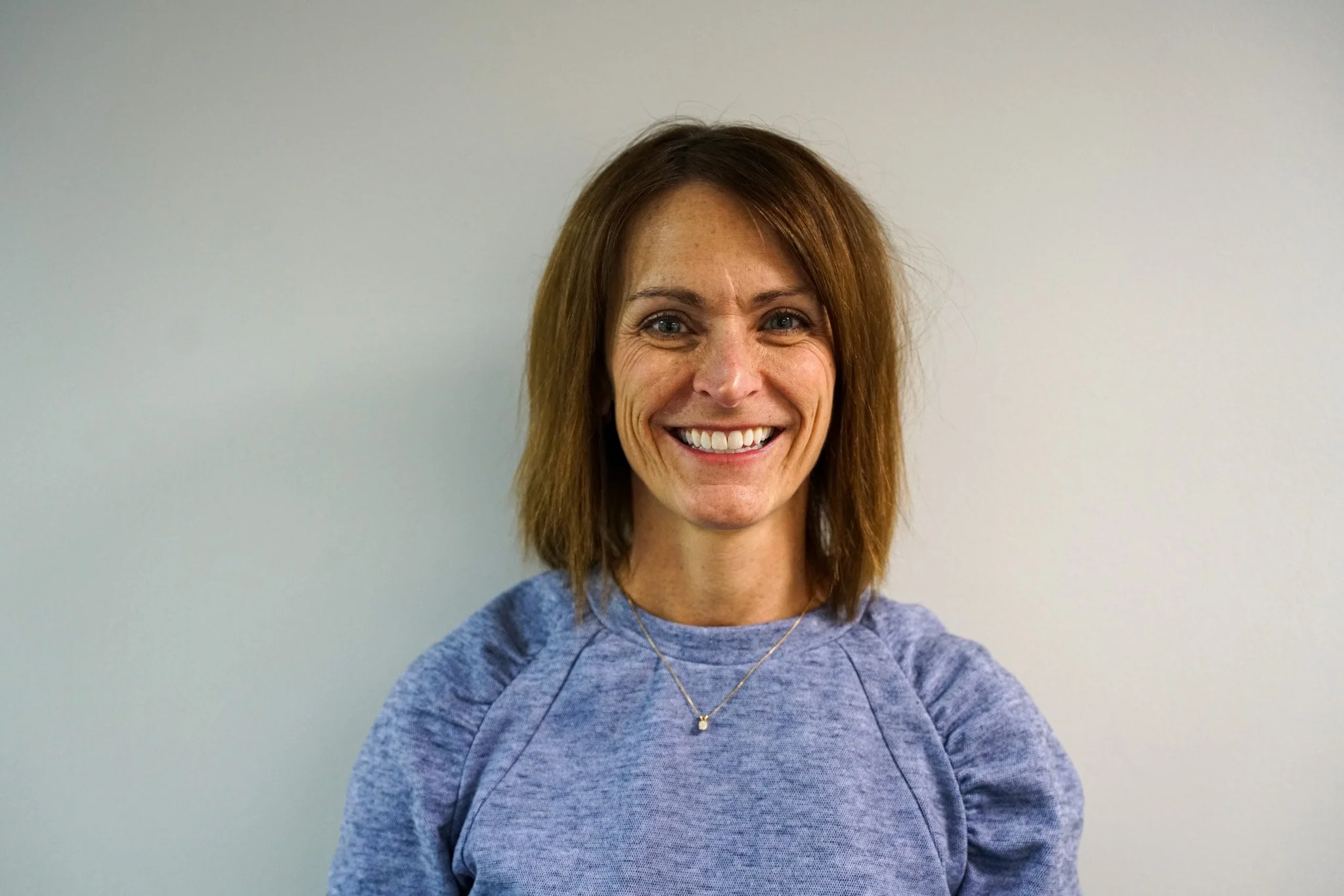 A woman with shoulder-length brown hair, smiling, wearing a light blue athletic top and a small gold necklace, standing against a plain light-colored wall.