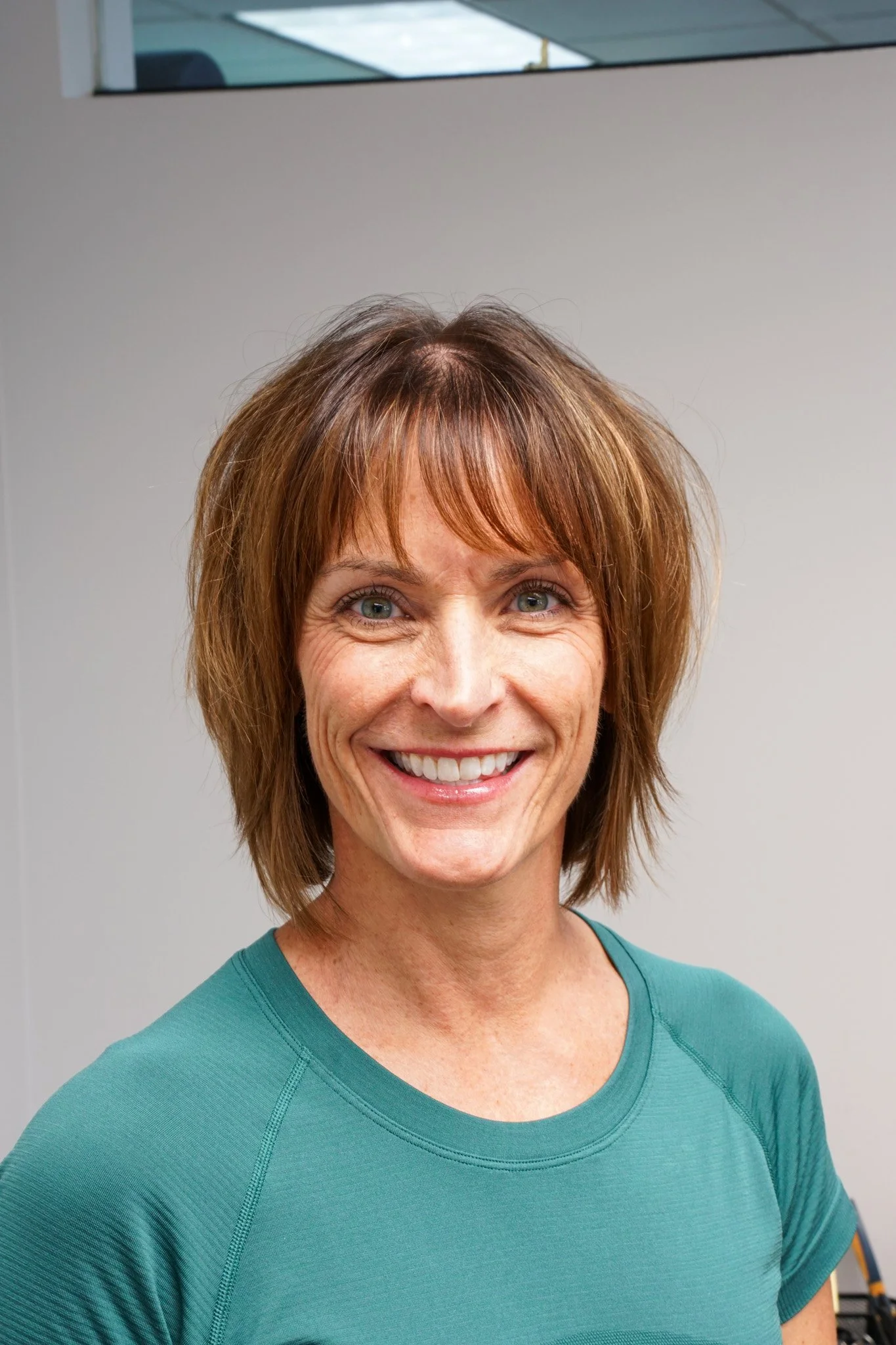 A woman with shoulder-length brown hair, smiling, wearing a light blue athletic top and a small gold necklace, standing against a plain light-colored wall.