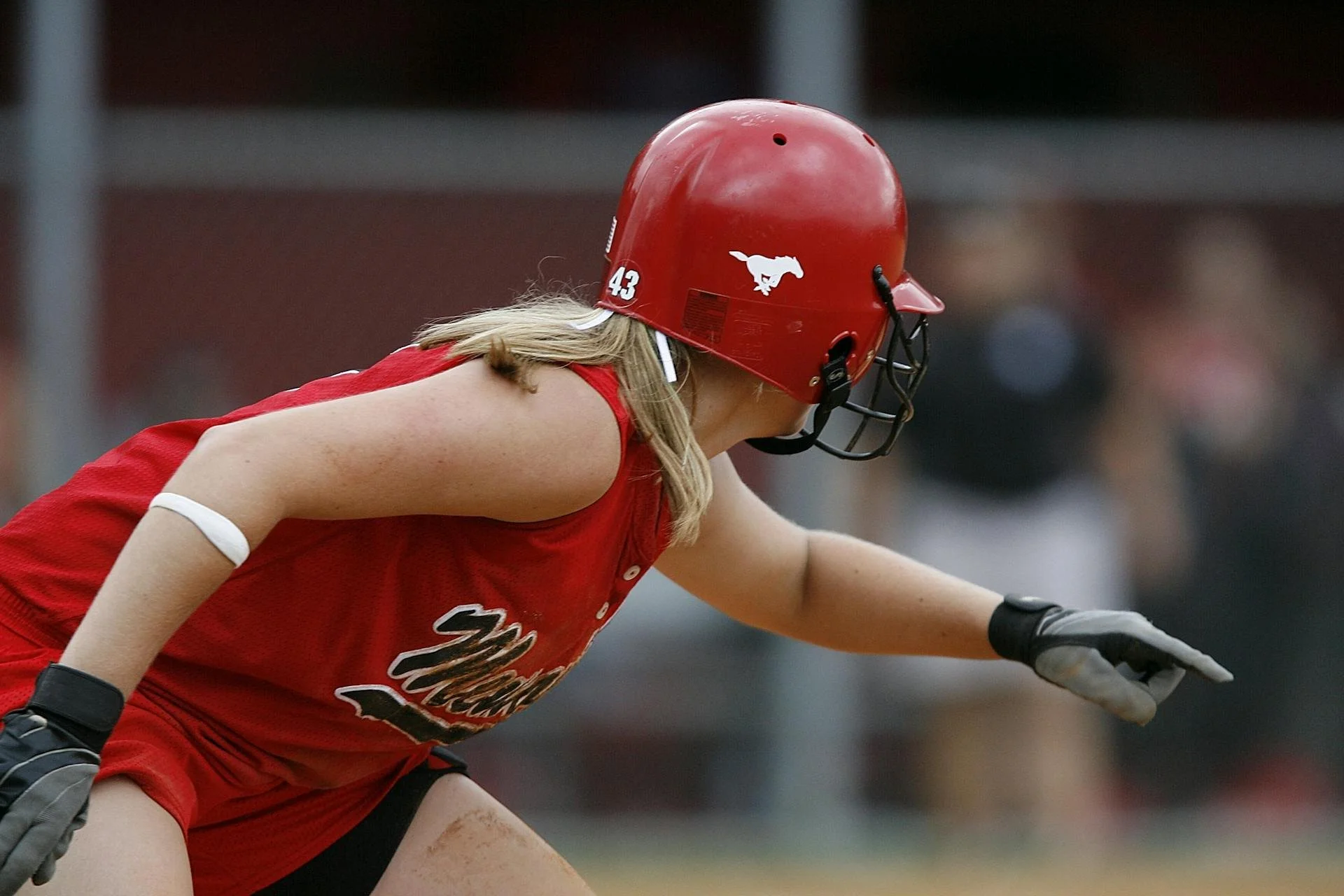Female athlete in a red sports uniform and helmet, crouching on a sports field, reaching with her right arm.