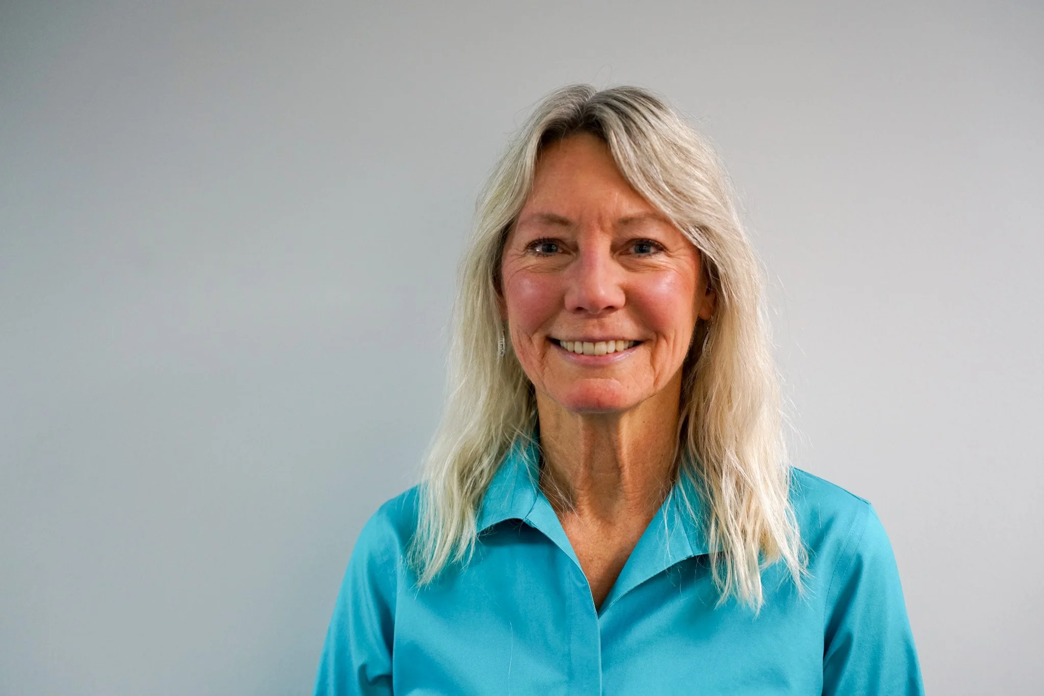 A smiling middle-aged woman with long, wavy, blonde hair wearing a turquoise shirt standing against a plain light gray background.