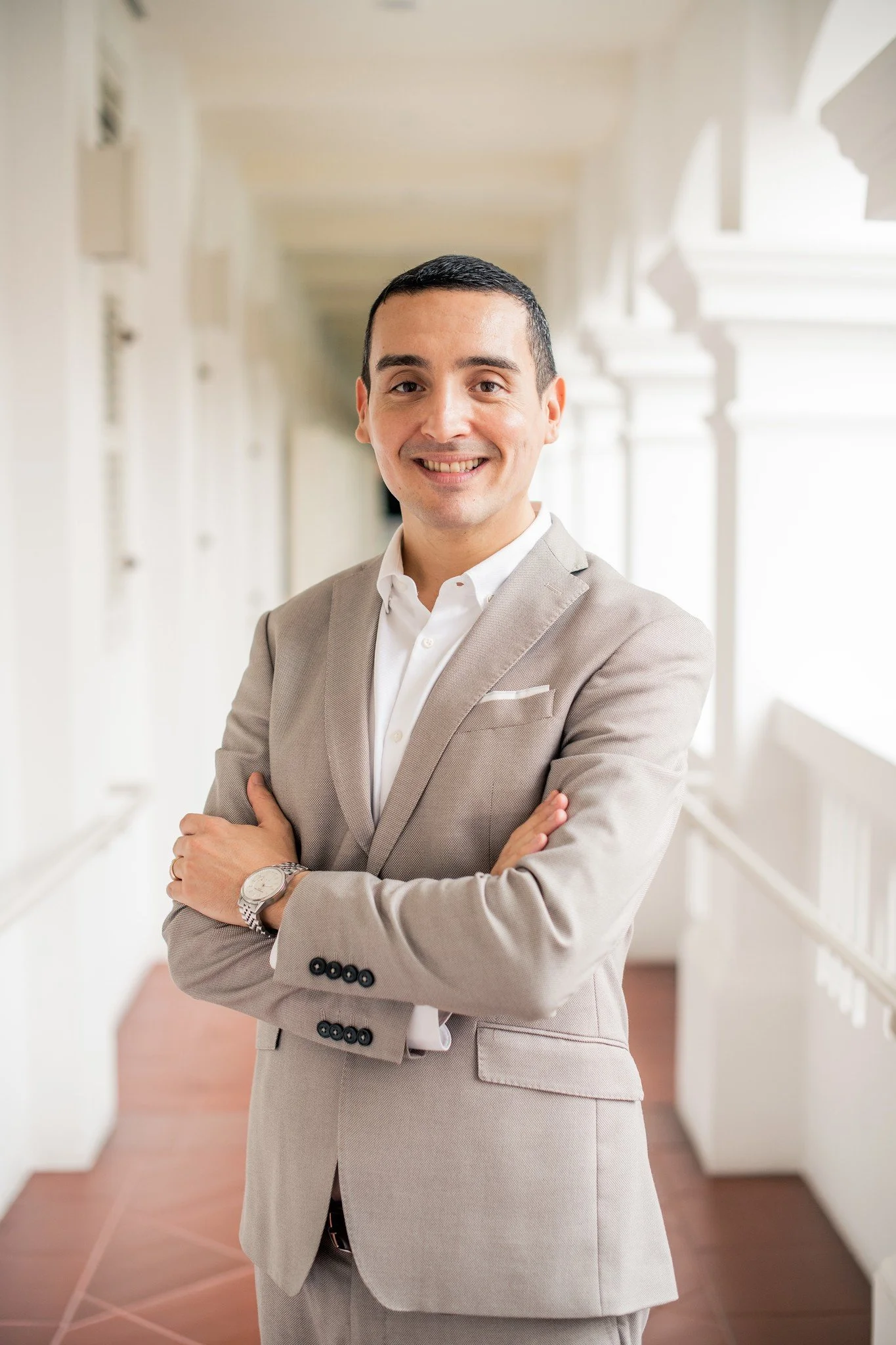 A smiling man in a beige suit with crossed arms standing in a bright, white hallway.