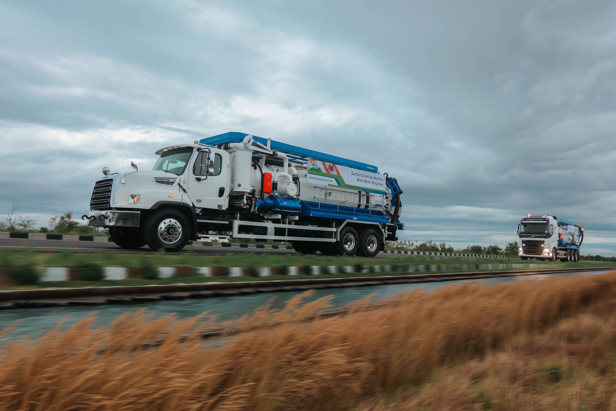 Two water recycling trucks driving on a highway under a cloudy sky, with blurred grass and road divider in the foreground.