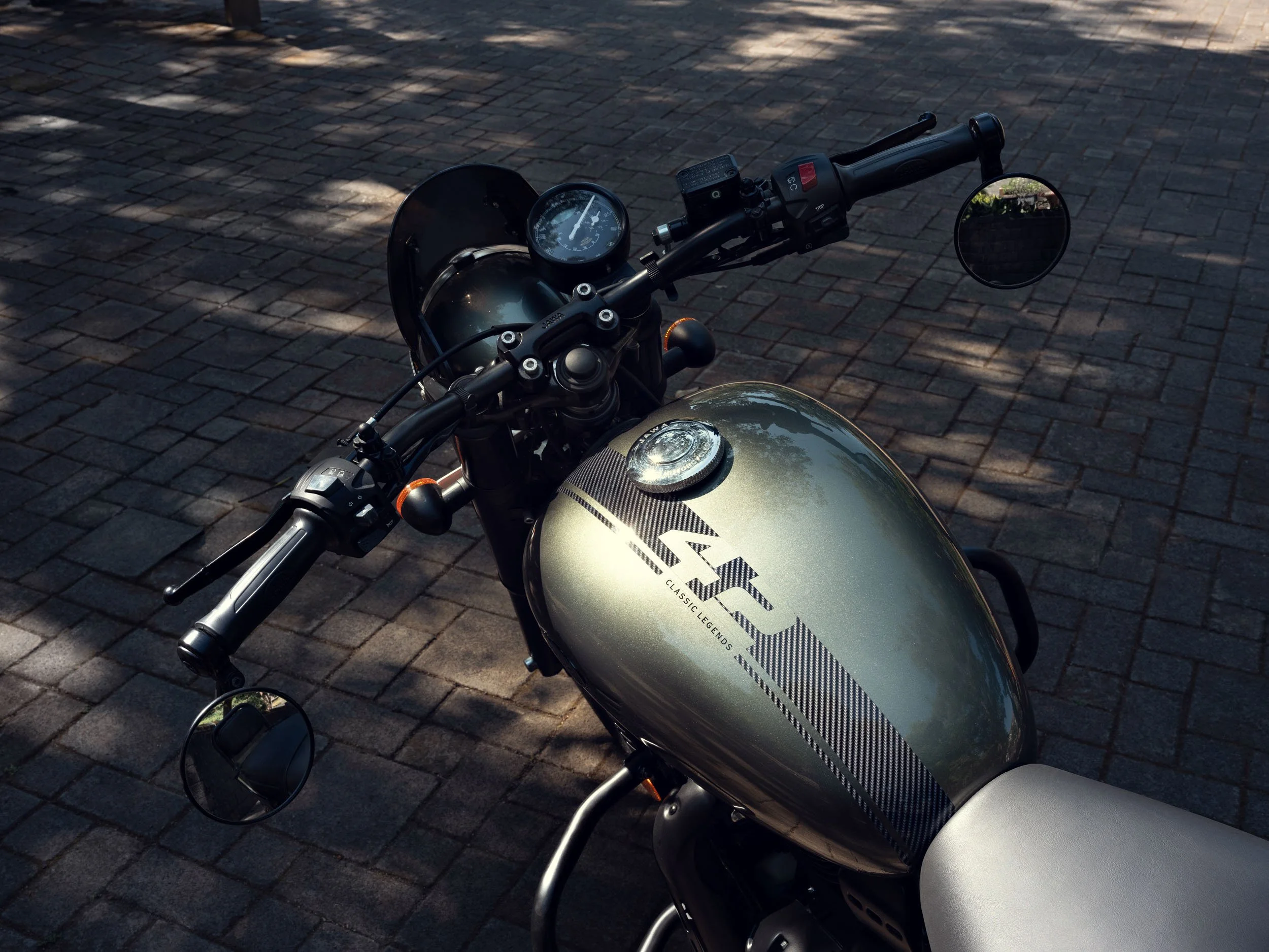A motorcycle with a metallic silver fuel tank and black accents parked on a brick-paved surface. The bike has round mirrors, a digital instrument panel, and a classic design with a modern touch.