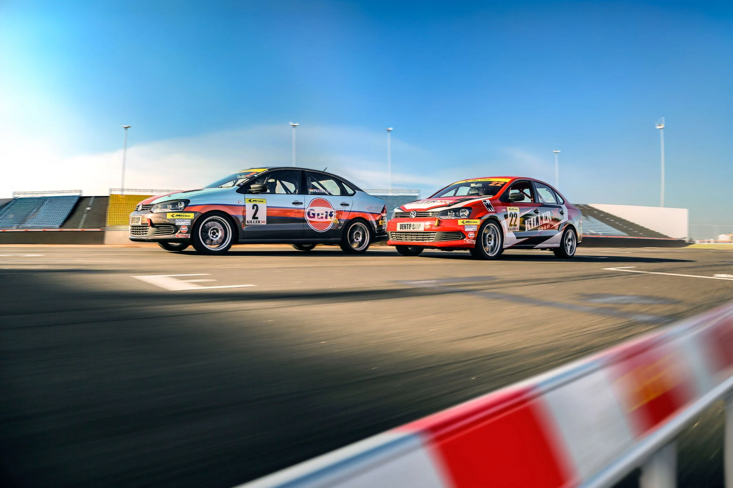 Two race cars on a track with a clear blue sky background. One is a silver and black car with the number 2, and the other is a red and white car with the number 22.