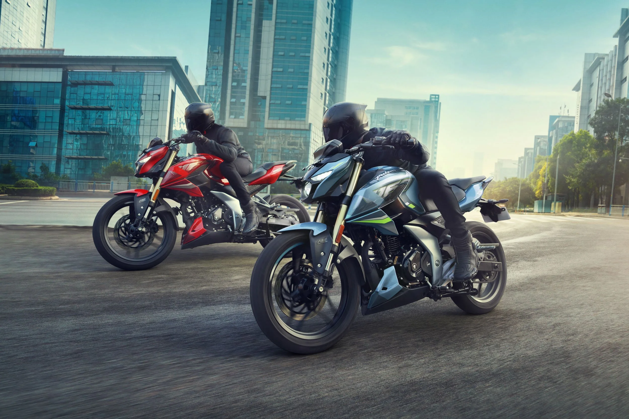 Two motorcyclists wearing black helmets and jackets riding sporty motorcycles on an urban street with modern glass buildings in the background.