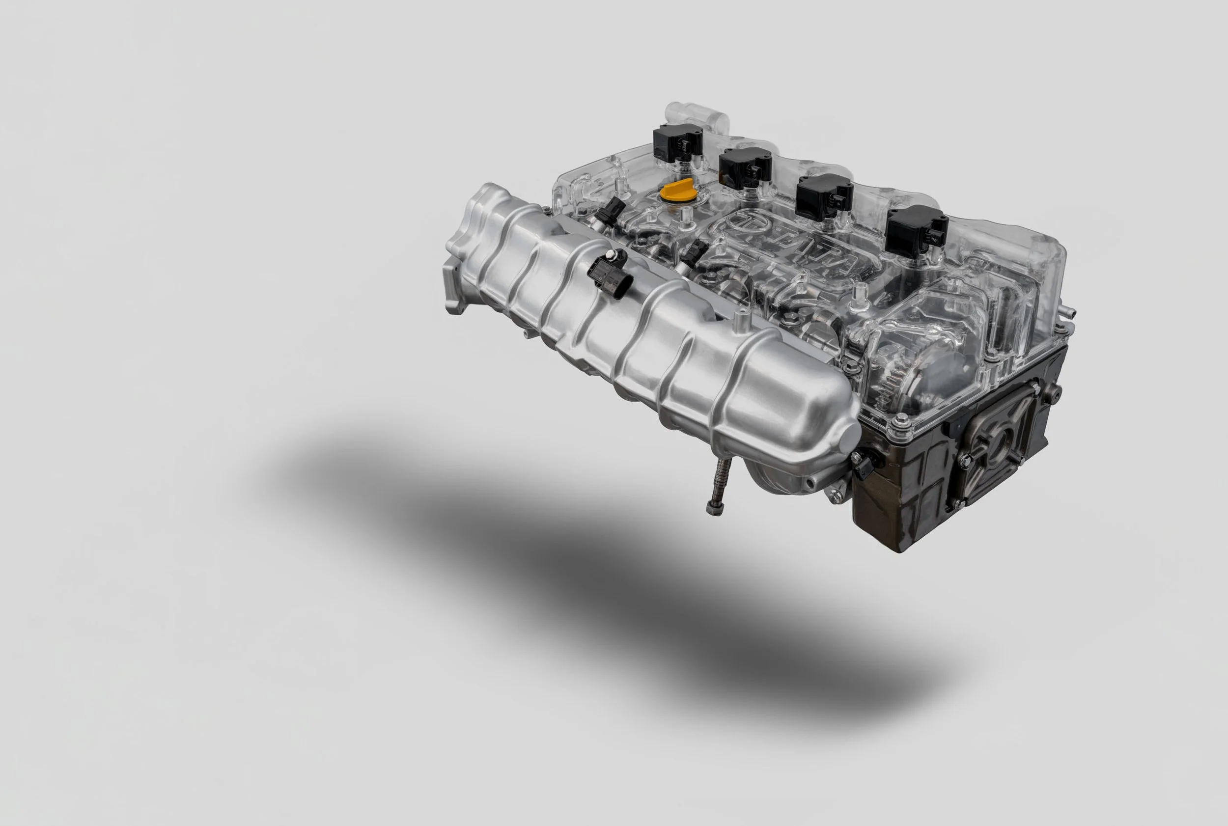 A modern automotive engine with a metallic intake manifold and transparent cover, showing internal components, against a plain gray background.