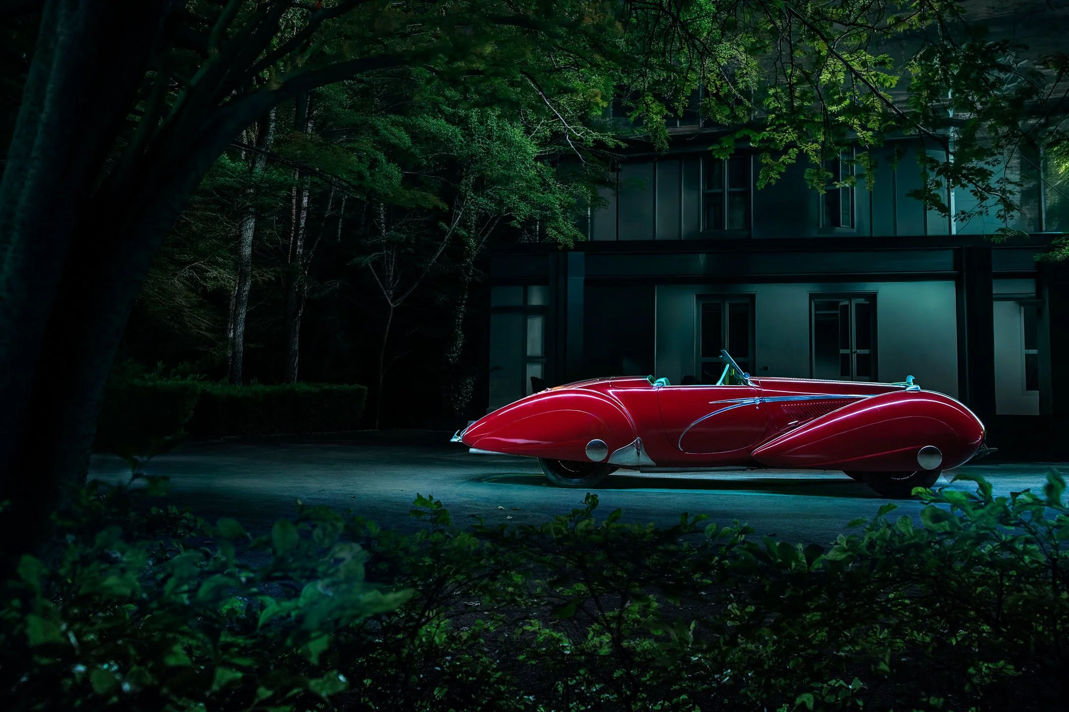 A vintage red car parked outside at night, surrounded by trees and a modern building in the background.