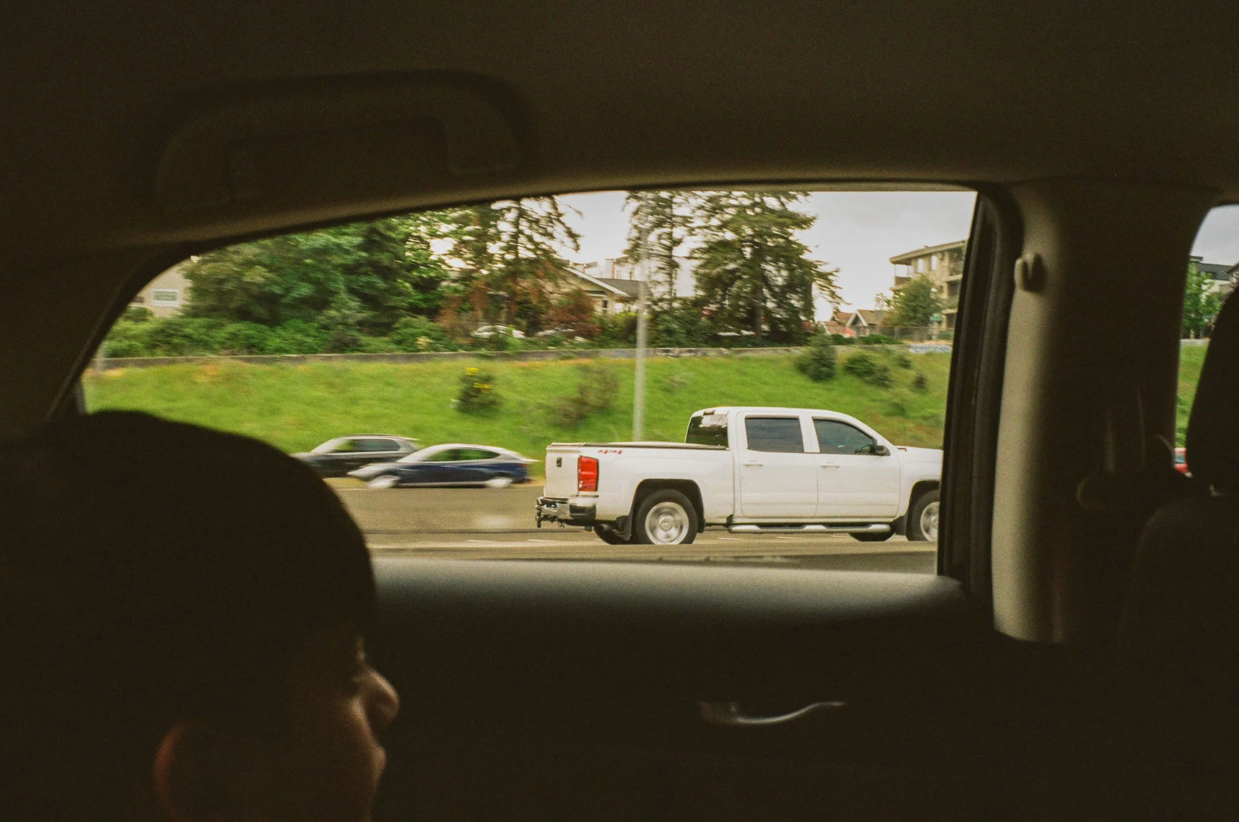 View from inside a vehicle looking through the rear window at a white pickup truck driving on a highway with trees and residential buildings in the background.