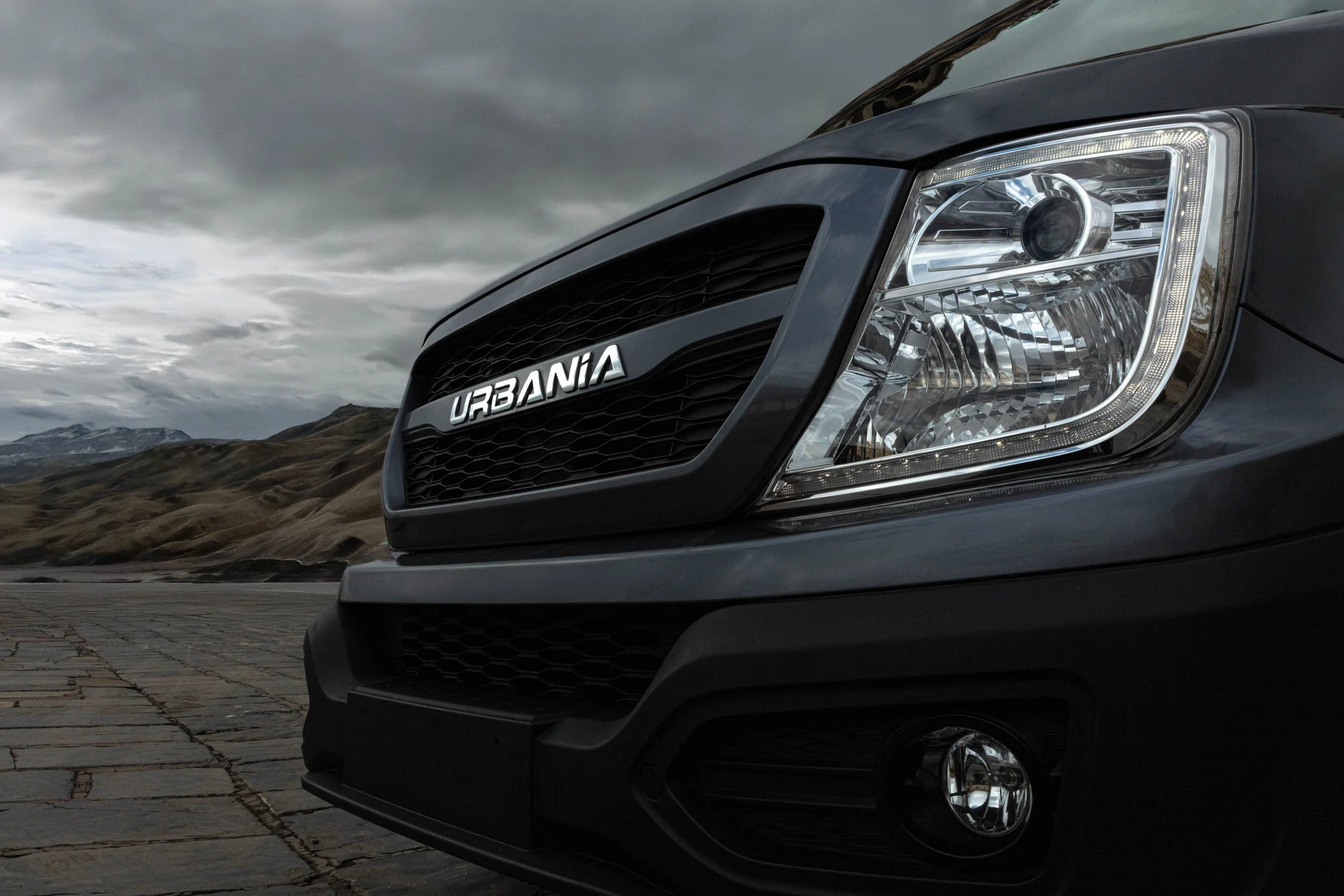 Close-up of the front of a black car with a chrome grille that says 'URBANIA,' parked on a cobblestone road with mountainous terrain and cloudy sky in the background.