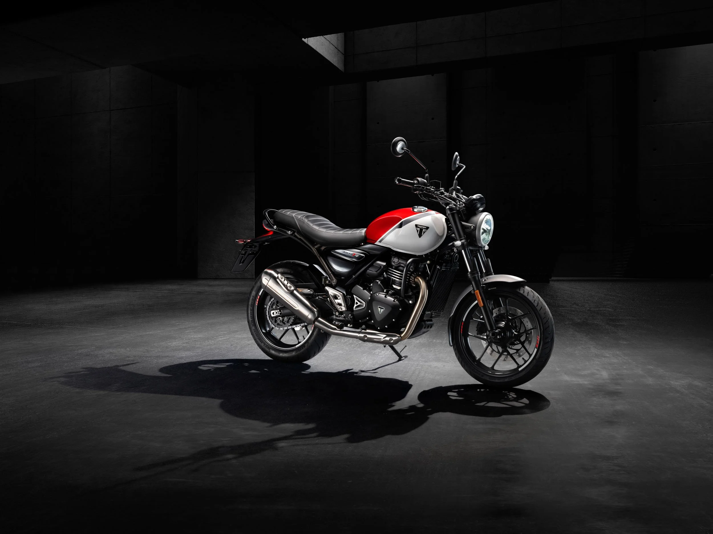 A modern motorcycle with a black and white body, red accents, and a chrome exhaust pipe, parked on a dark, concrete surface with a shadow beneath it, in a dimly lit industrial setting.