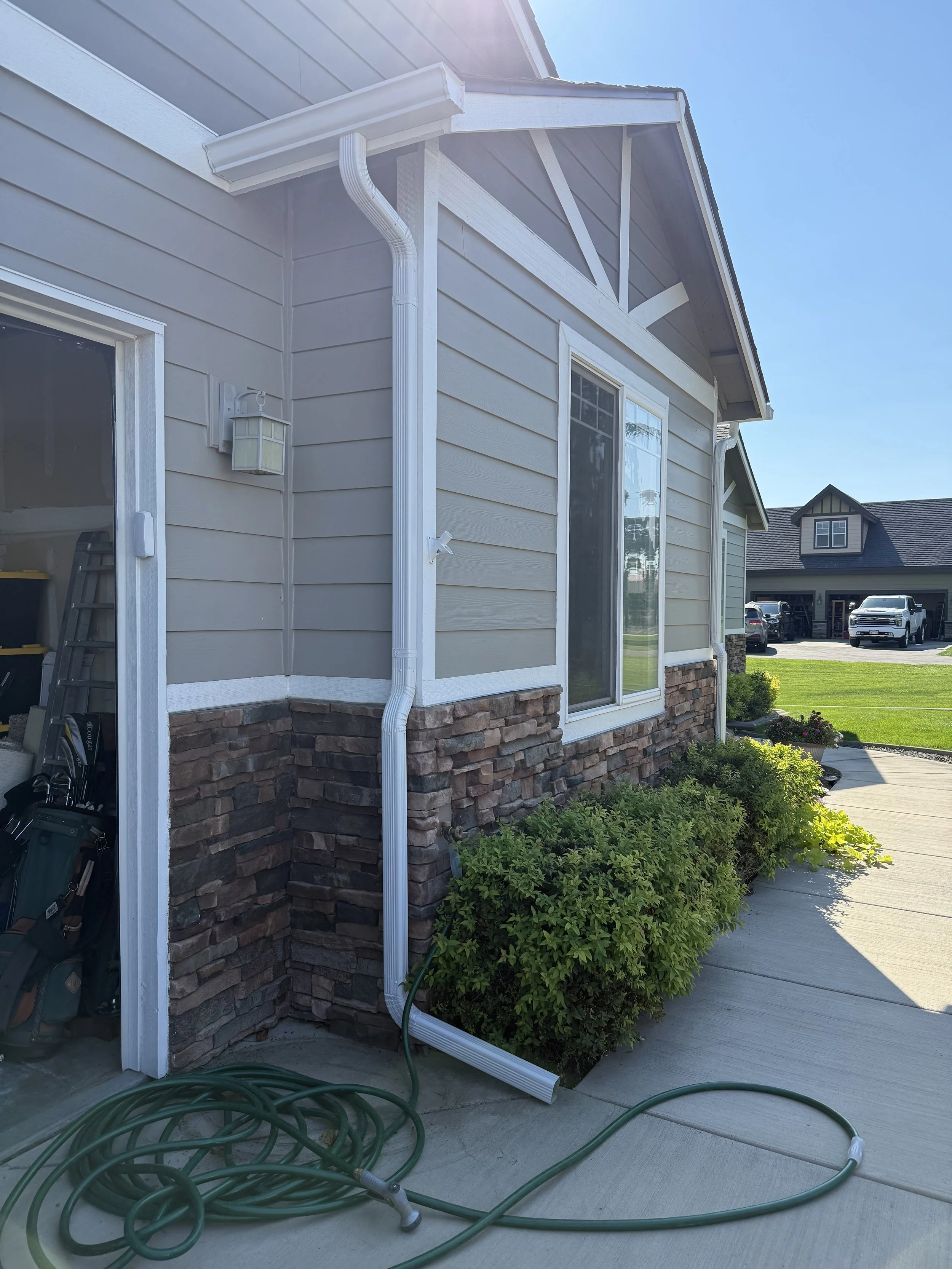 Side of a house with beige siding and a stone foundation, showing a downspout extender, garden hoses on the concrete driveway, a window, and bushes, with a view of neighboring houses and a clear sky in the background.
