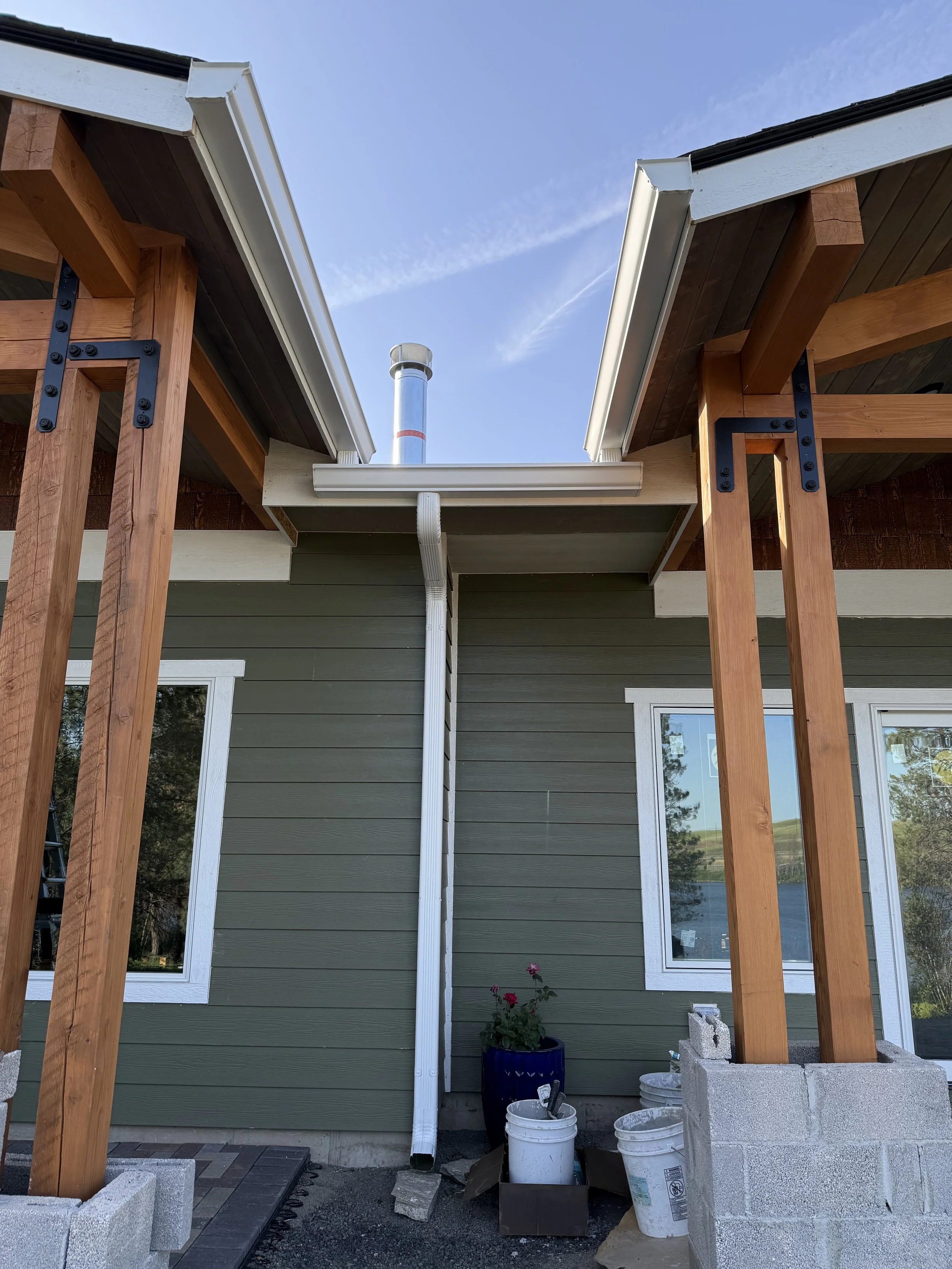 Partially constructed house exterior with green siding, white-trimmed windows, wooden support beams, and a new gutter system under construction. Some construction materials and tools are visible at the base.