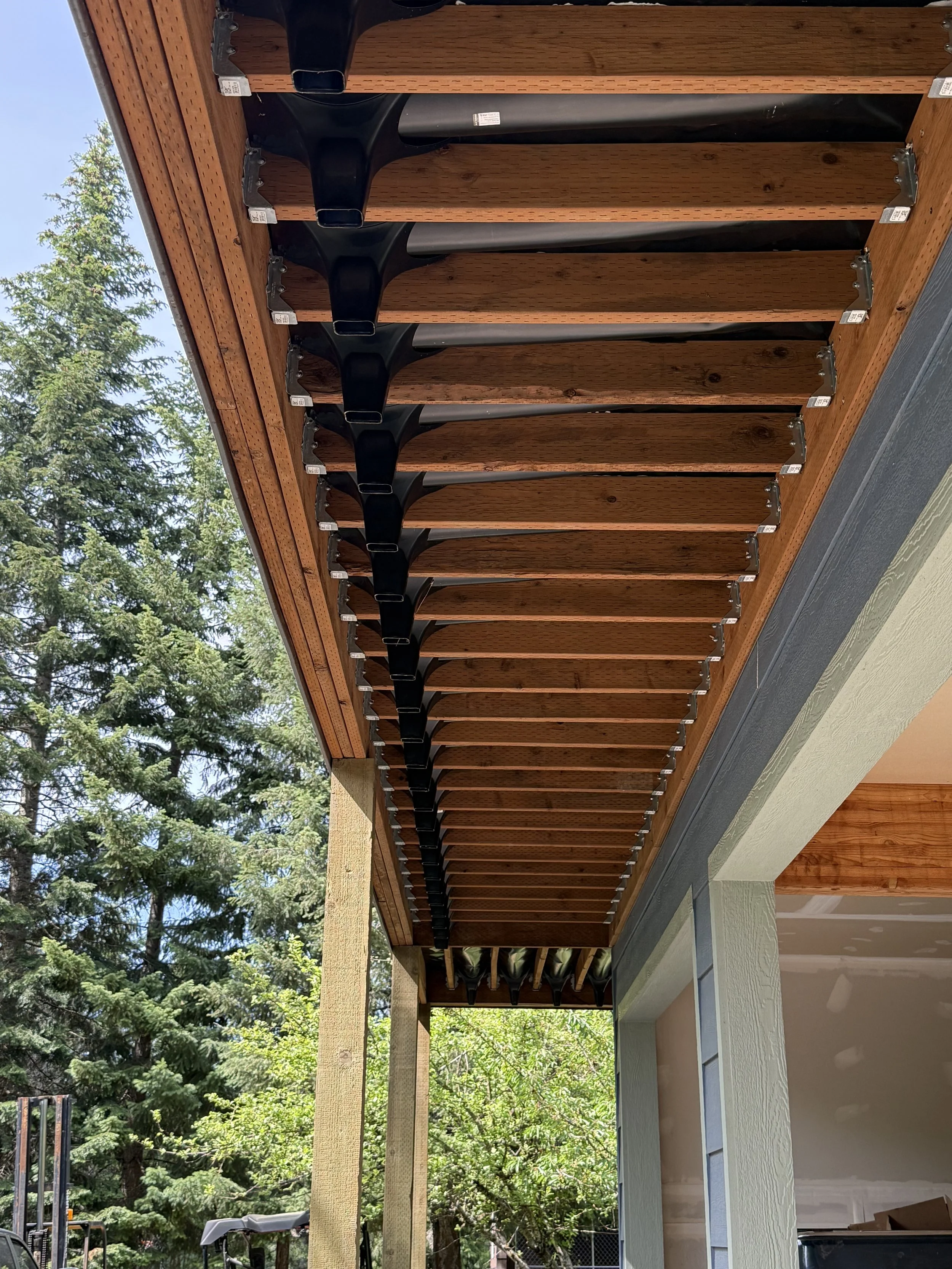 Under construction of a wooden porch roof with black steel brackets supporting the overhang, with a backdrop of trees and blue sky.