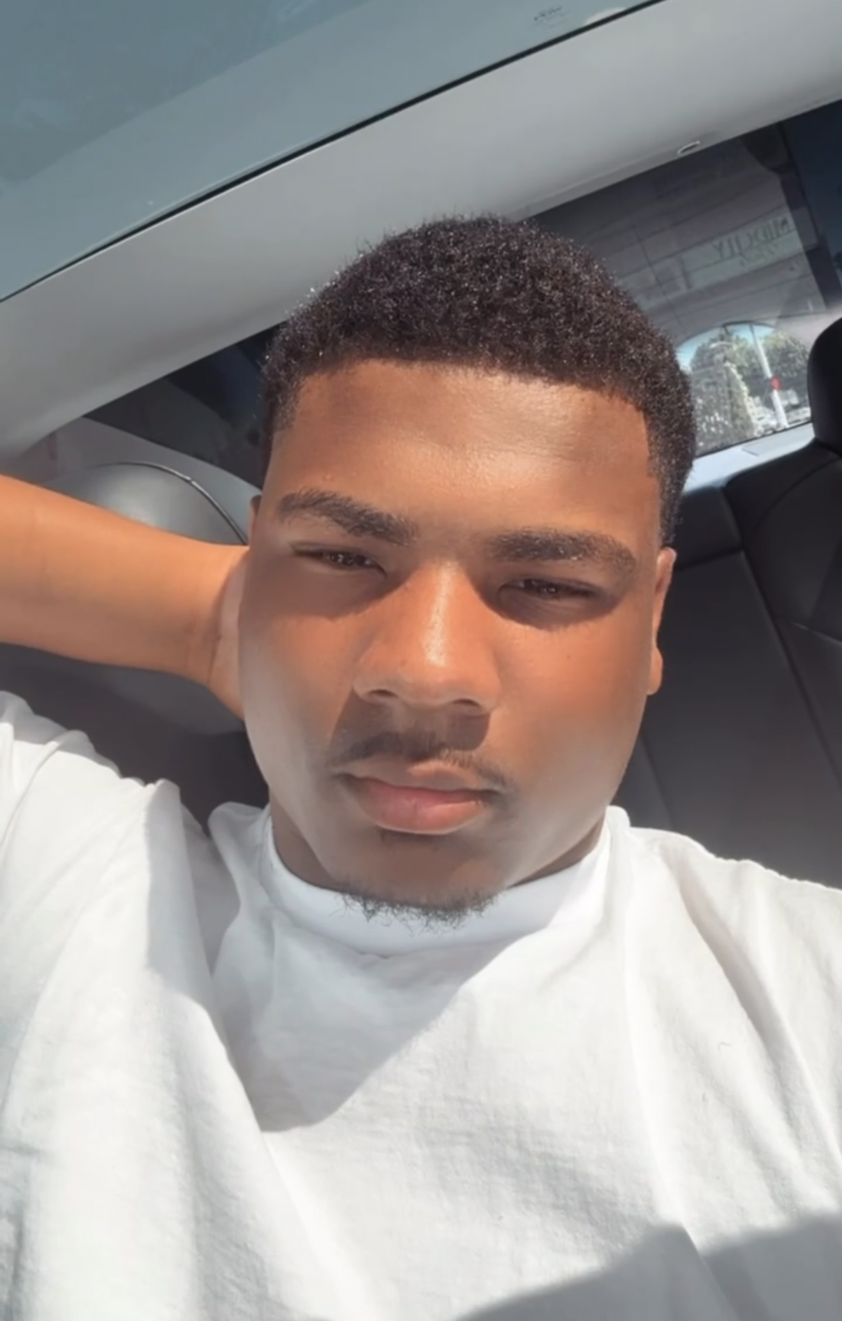 A young man taking a selfie inside a car, lying down with one arm behind his head, sunlight illuminating his face, and trees visible through the window.