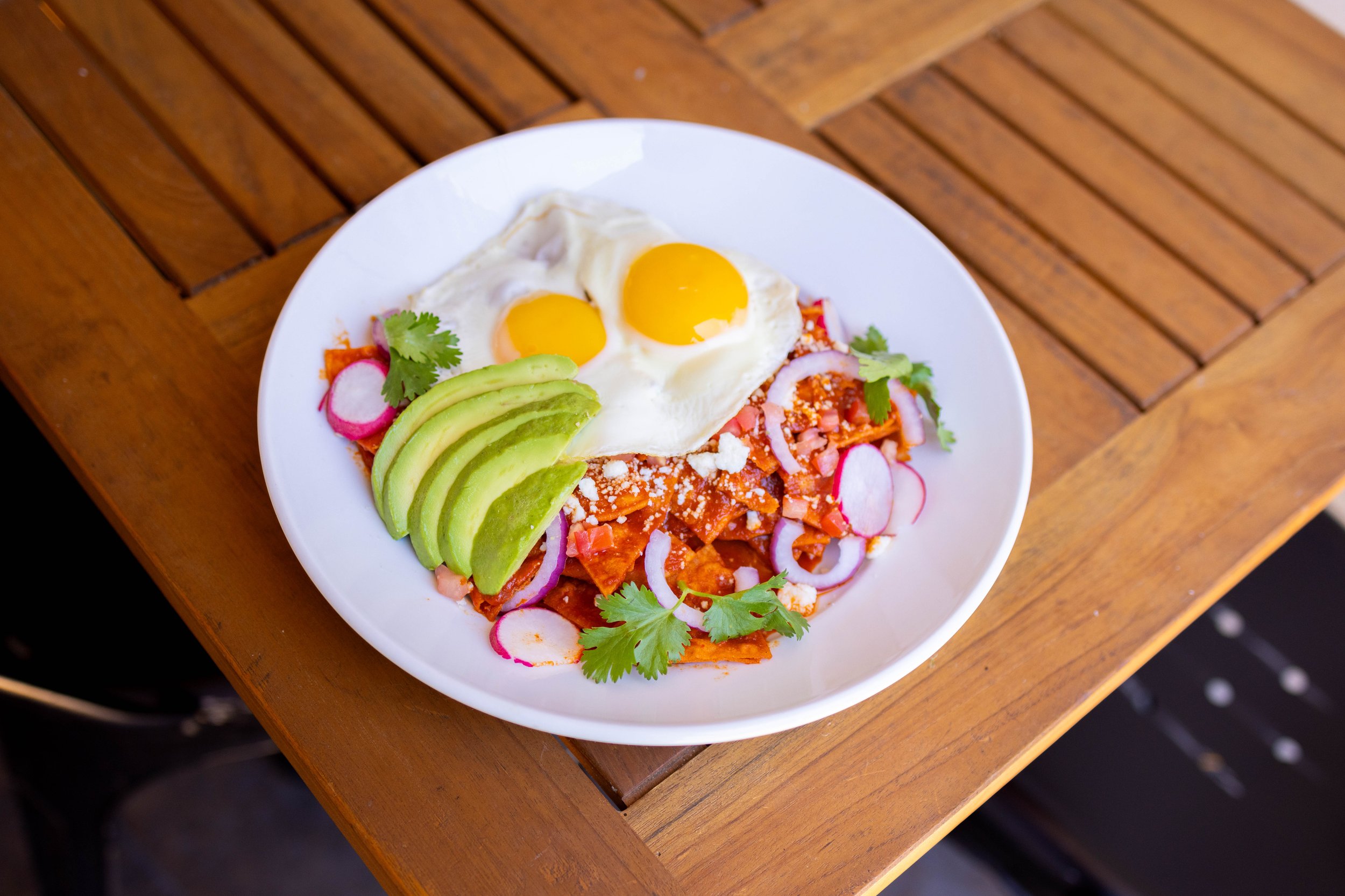 Plate of chilaquiles topped with two fried eggs, sliced avocado, radishes, onion slices, cilantro, crumbled cheese, and red sauce, on a wooden table.