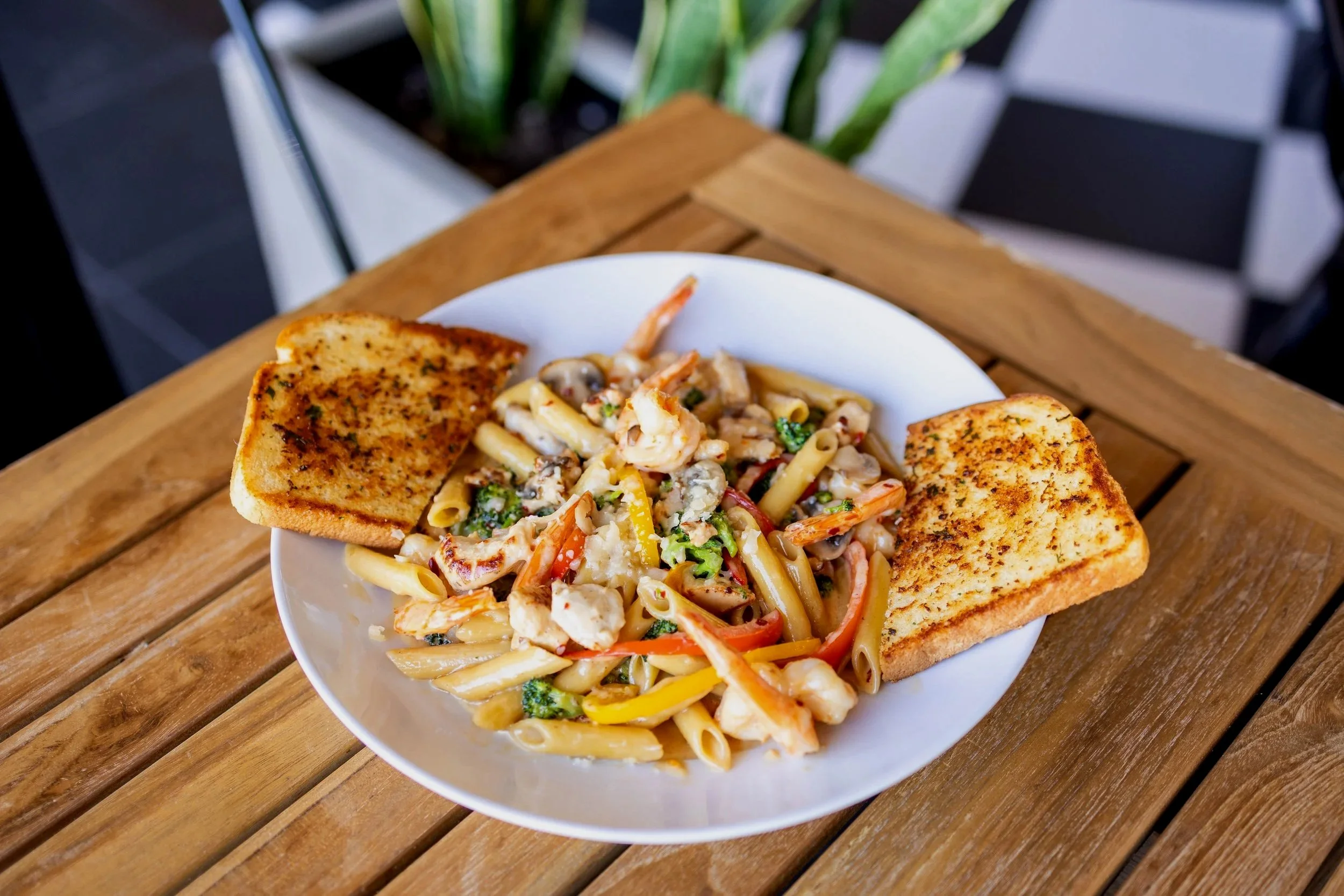 A white bowl with pasta salad containing colorful vegetables and shrimp, accompanied by two slices of toasted bread on a wooden table.