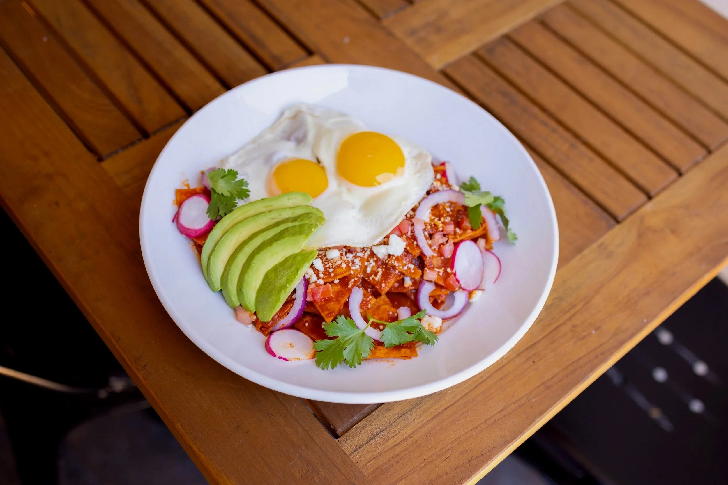 Plate of chilaquiles topped with two sunny-side-up eggs, sliced avocado, radish slices, cilantro, and crumbled cheese.