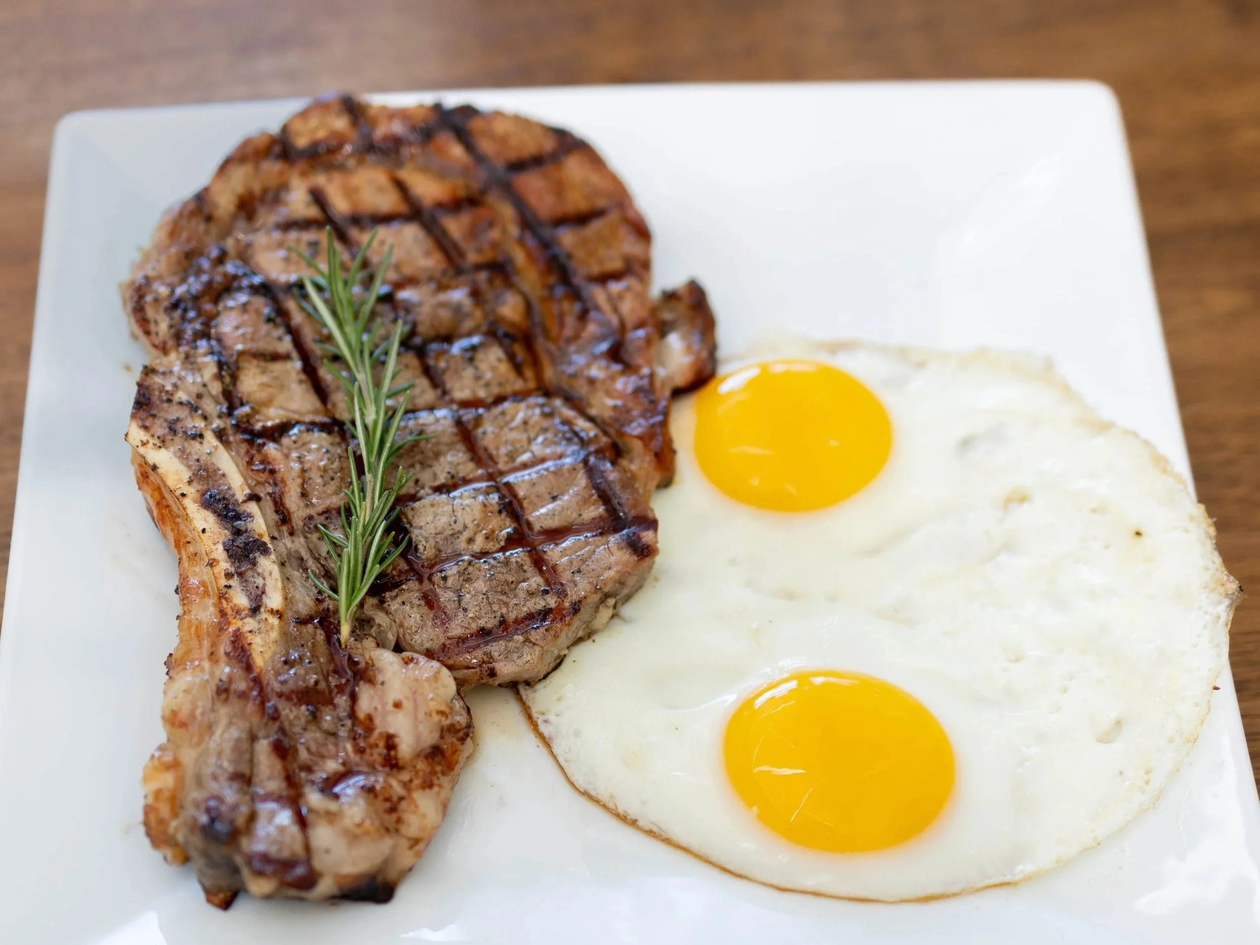 Steak with grill marks, two sunny-side-up eggs, and a sprig of rosemary on a white square plate.