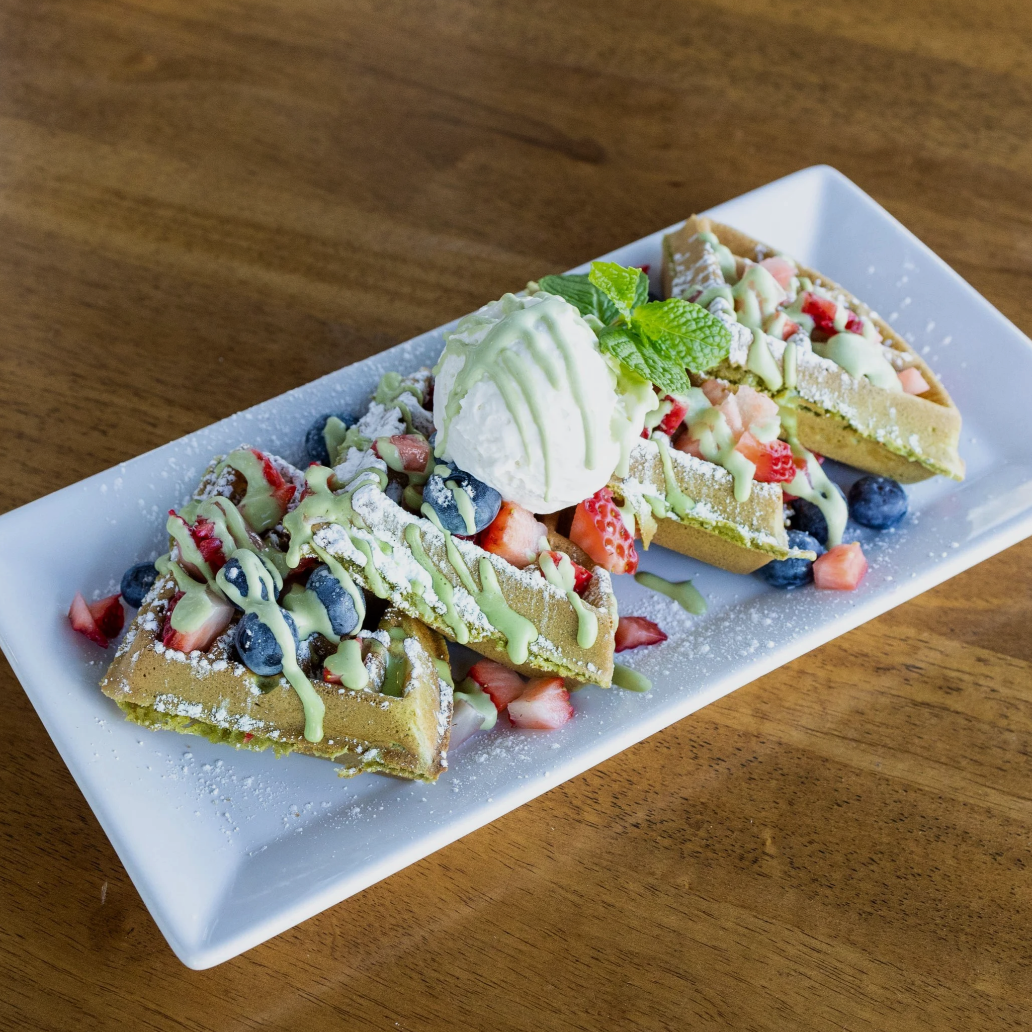 Three waffle pieces topped with mixed berries, white chocolate drizzle, powdered sugar, a scoop of vanilla ice cream, and a mint leaf on a white rectangular plate.