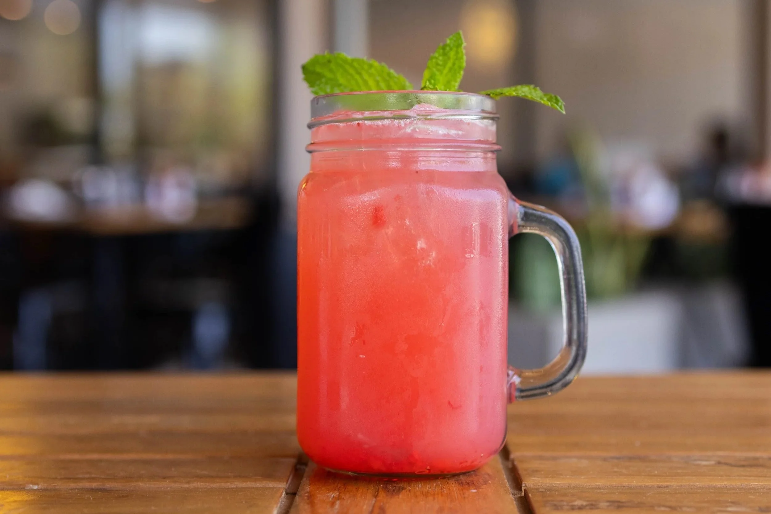 A pink fruit smoothie in a glass mason jar with a handle, garnished with a sprig of fresh mint, on a wooden table.