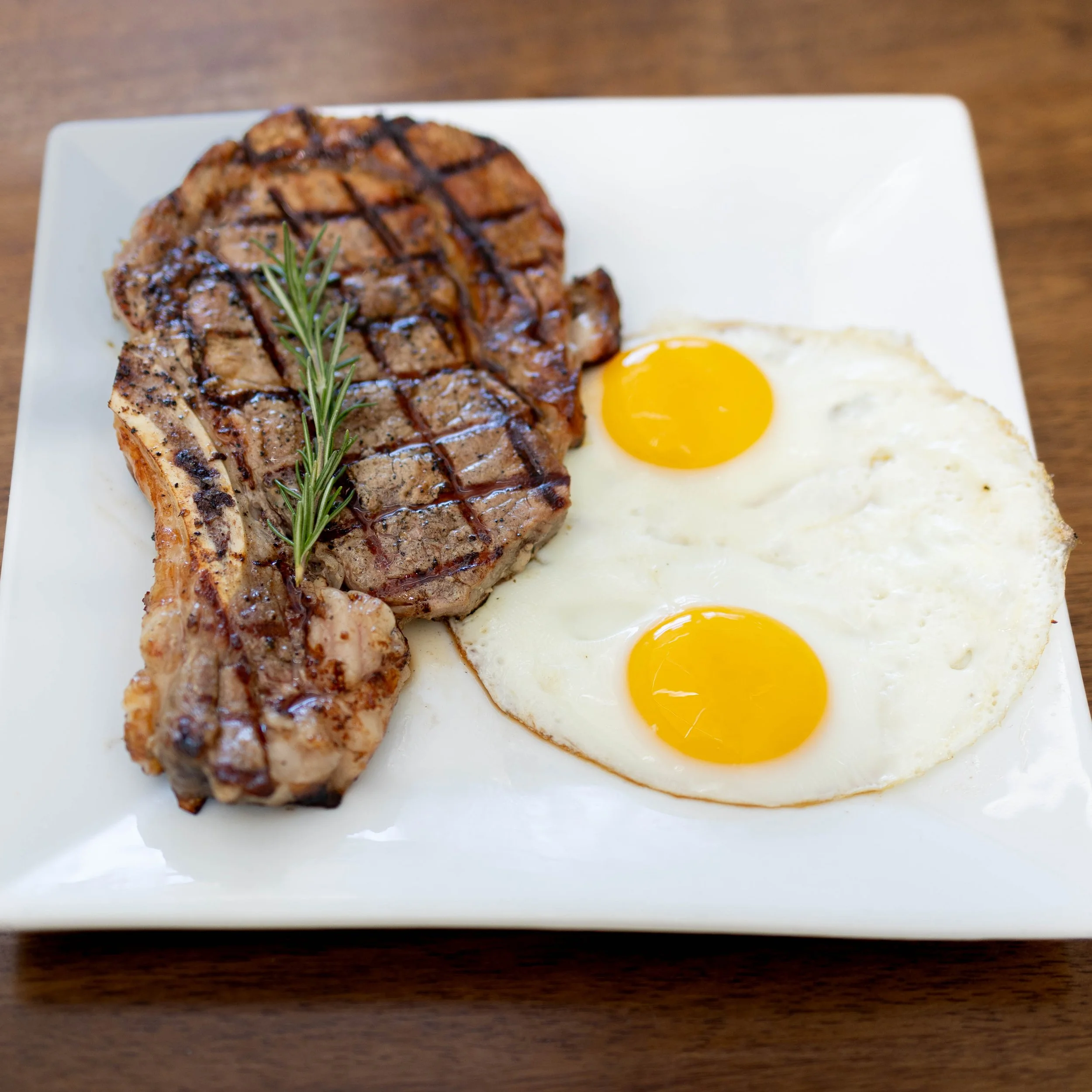 Steak with grill marks, grilled chicken, sunny side up eggs on a white square plate garnished with a sprig of rosemary.