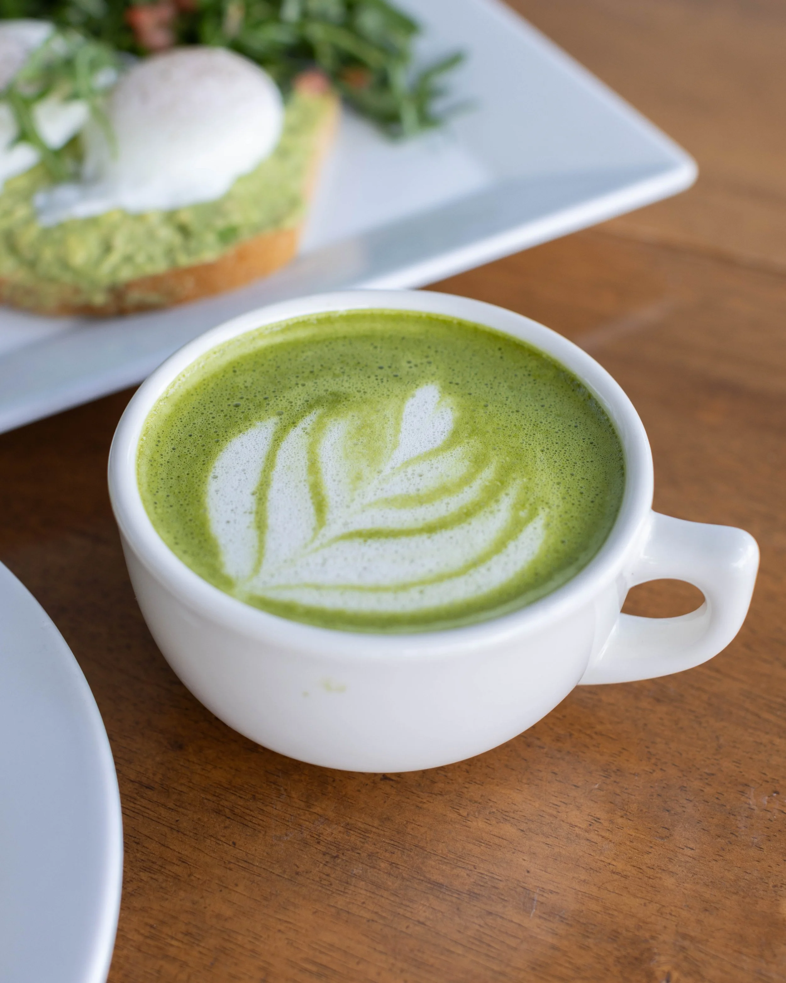 A white cup of matcha latte with latte art, placed on a wooden table, with an avocado toast with poached egg and greens in the background.
