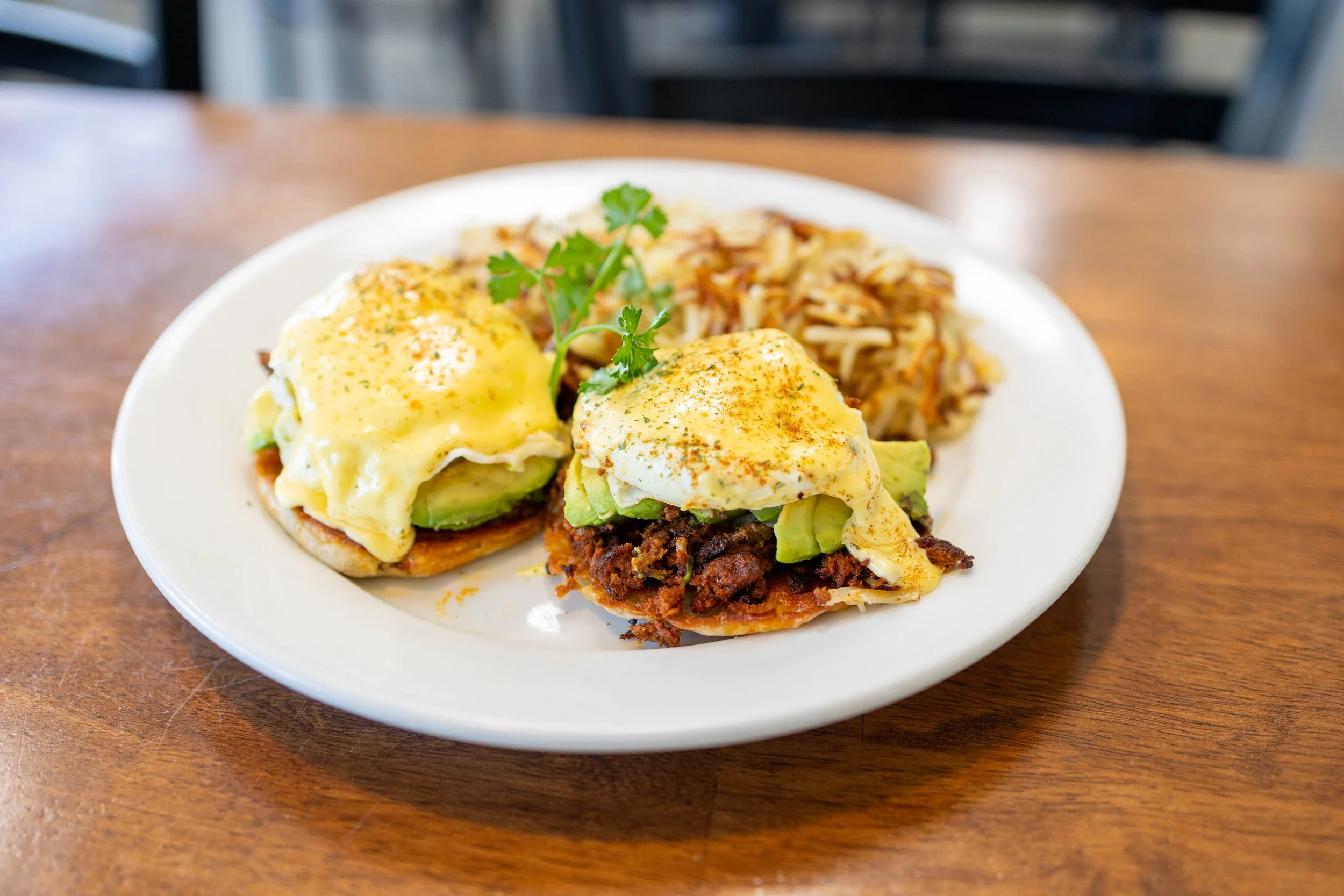 Plate of eggs benedict with avocado and bacon, alongside seasoned shredded hash browns on a white plate.
