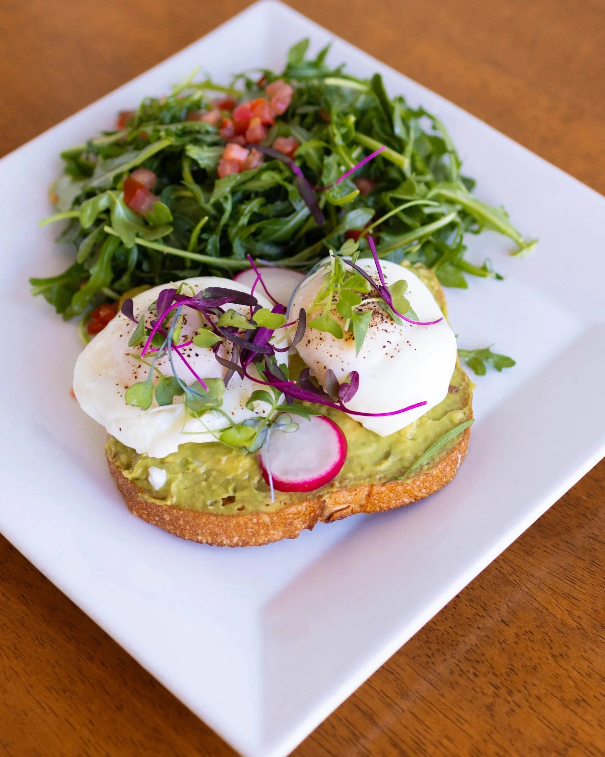 Poached eggs on avocado toast with microgreens, radish slices, and a side salad of leafy greens with tomatoes on a white plate.