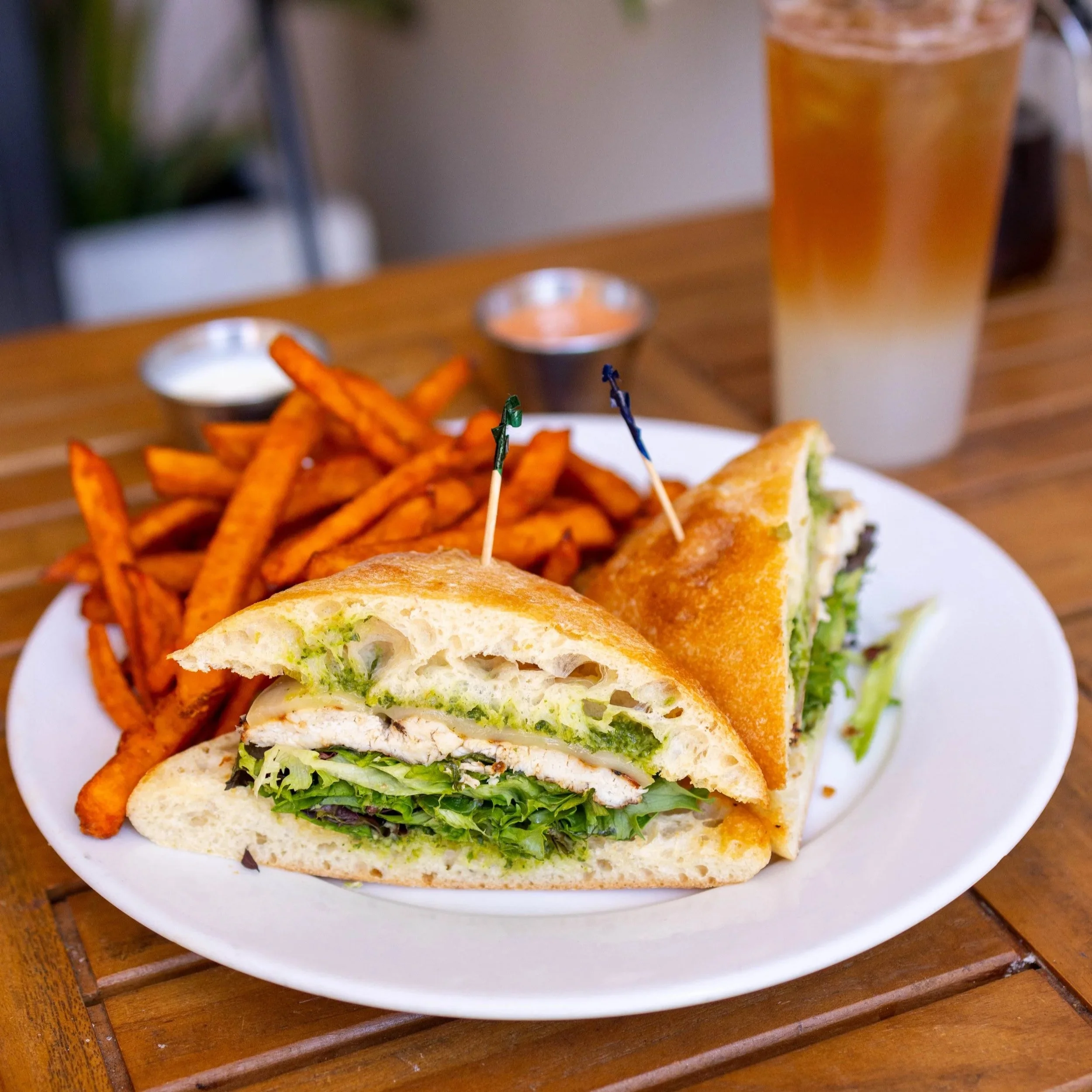 A plate with a chicken club sandwich cut in half, with lettuce, tomato, and green sauce, served with sweet potato fries, two small containers of dipping sauce, and a glass of iced tea in the background.