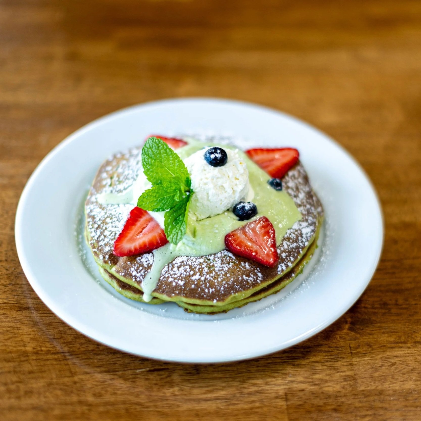 Pancakes topped with strawberries, blueberries, mint leaves, whipped cream, and powdered sugar, with a green sauce drizzled over