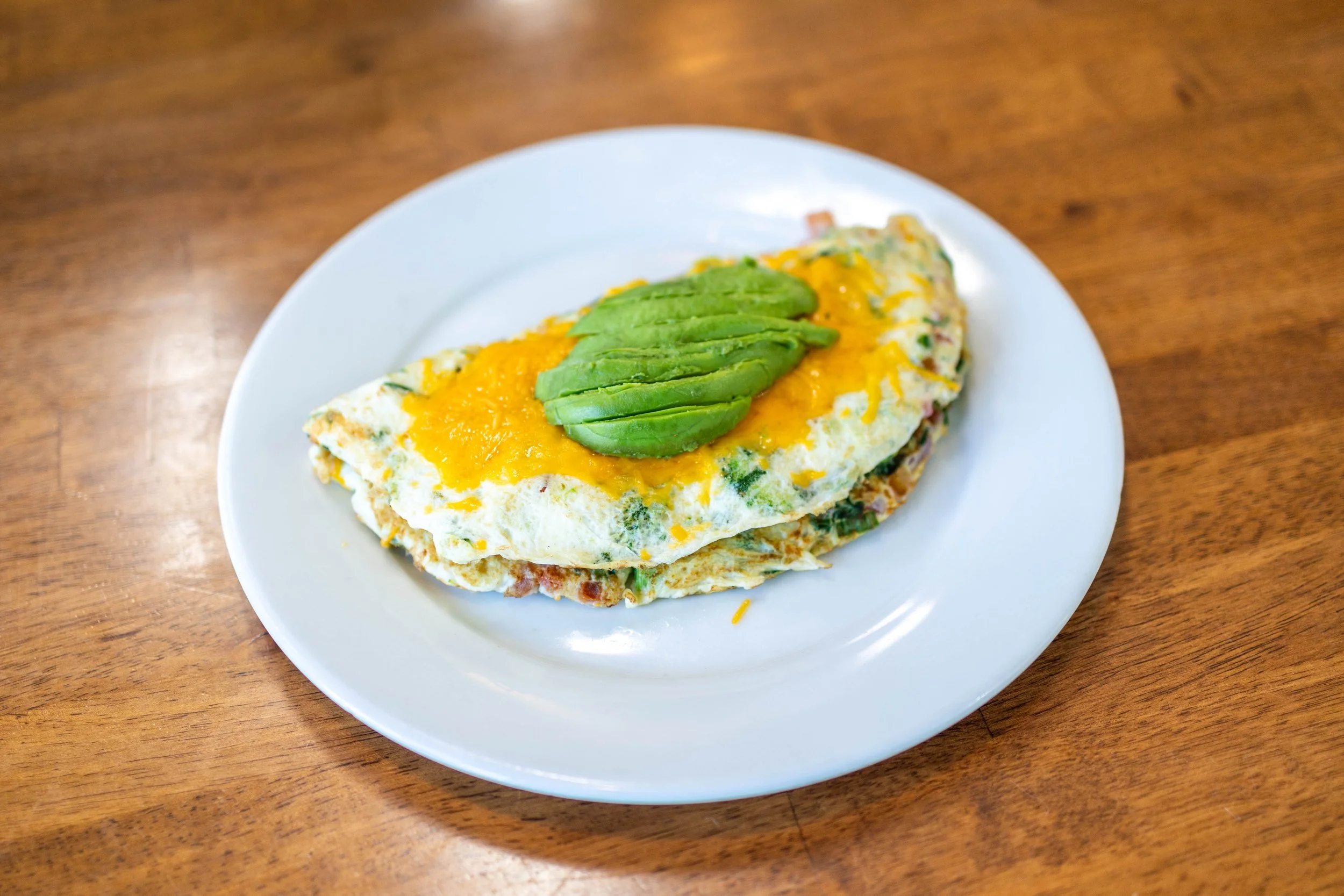 Omelette topped with shredded cheddar cheese and sliced avocado served on a white plate.