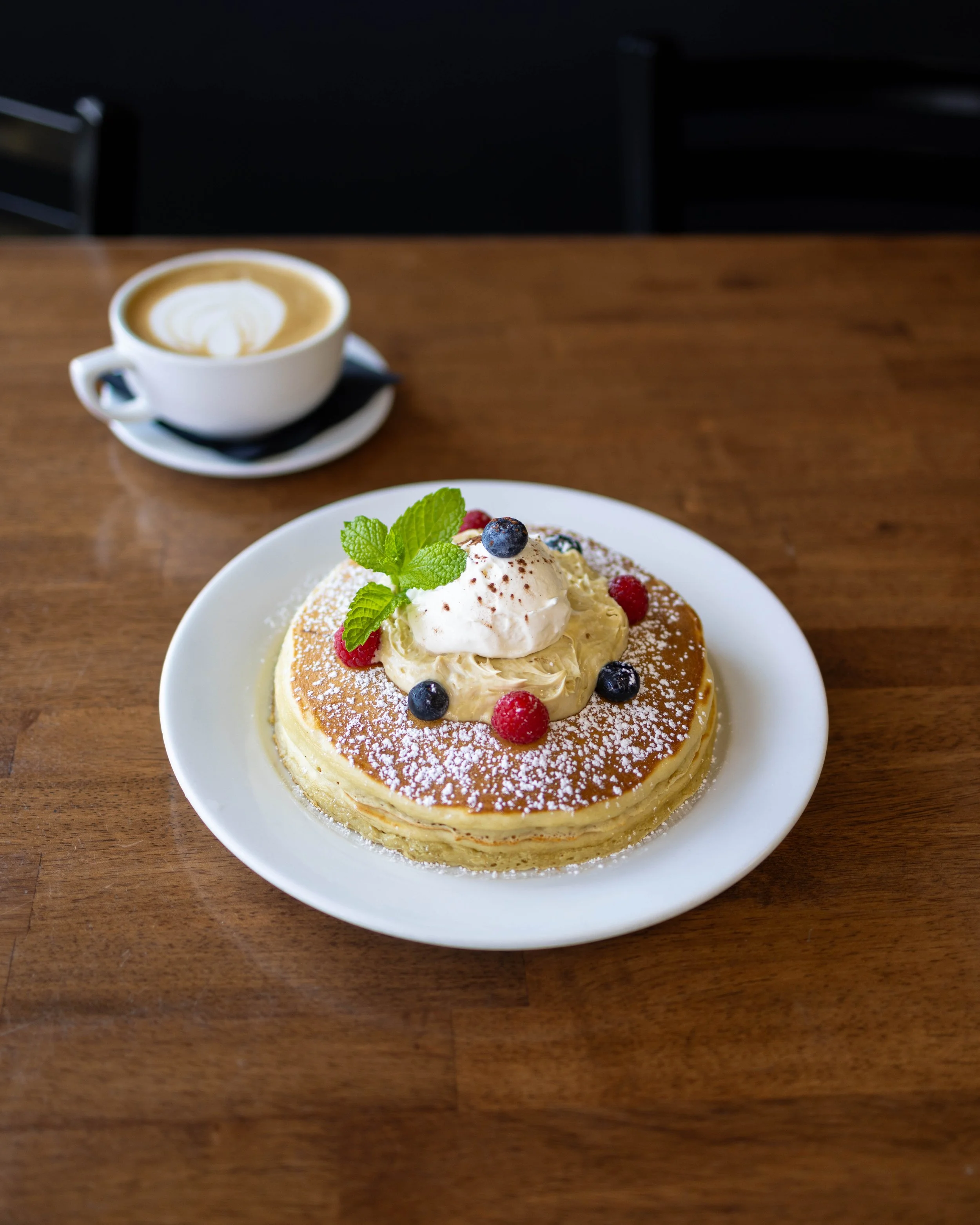 A plate of pancakes topped with whipped cream, berries, mint leaves, and powdered sugar, with a cup of coffee in the background.