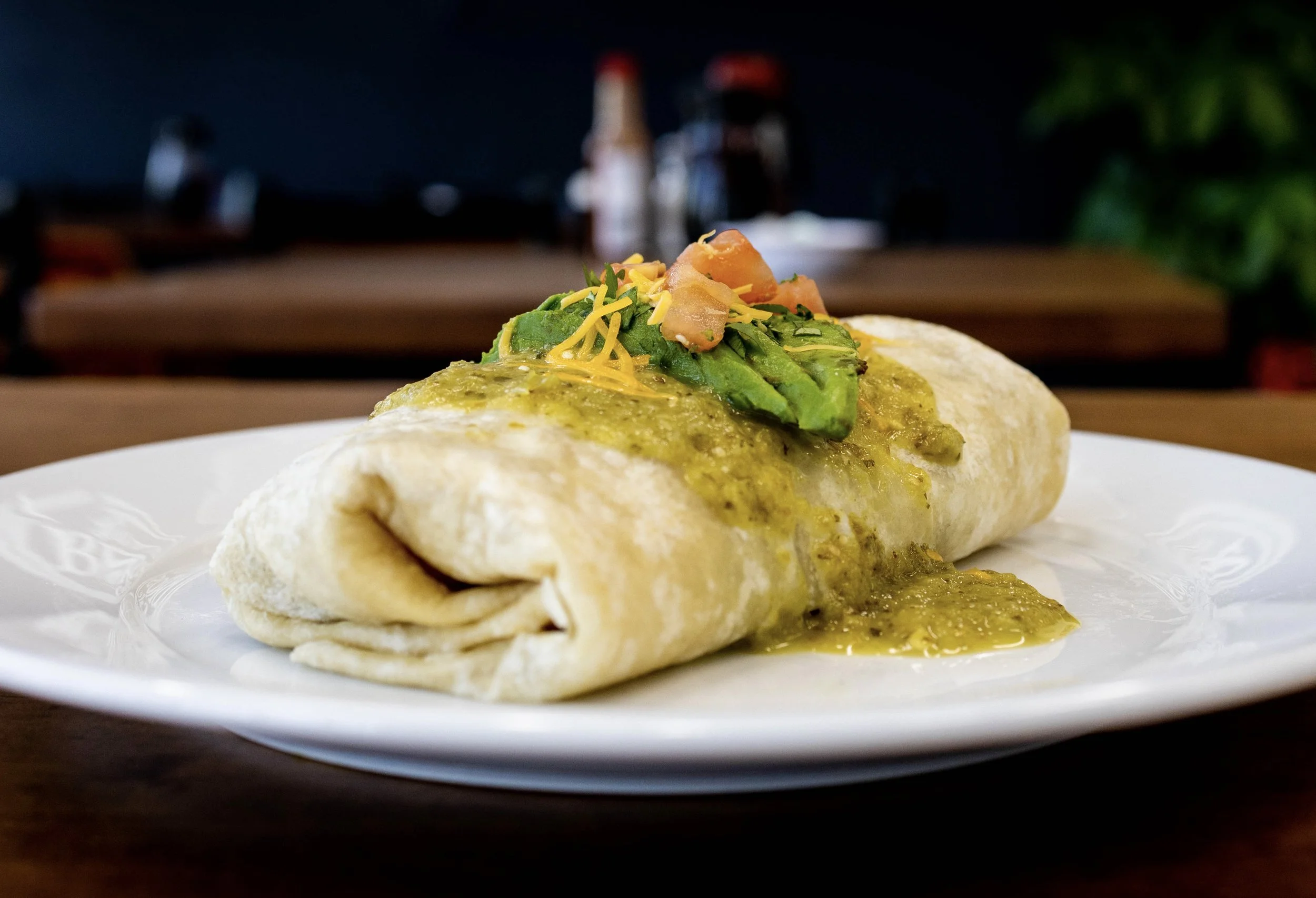 A plate with an stuffed burrito topped with avocado slices, shredded cheese, and salsa in a restaurant setting.