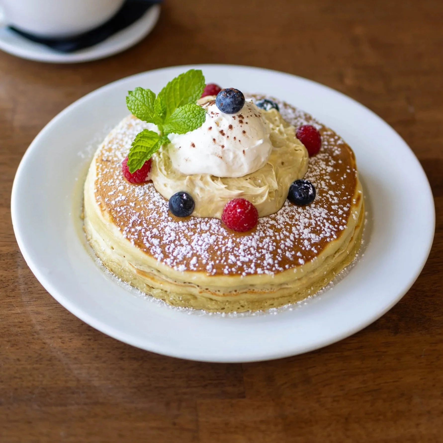 Pancakes topped with whipped cream, fresh blueberries, raspberries, mint leaves, and powdered sugar on a white plate.