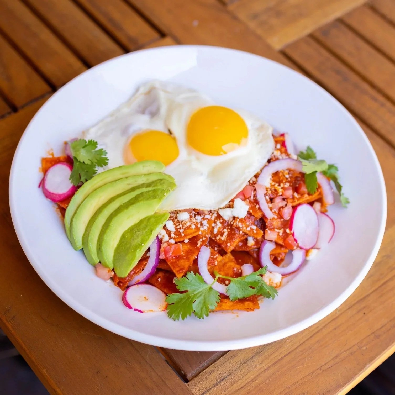 A plate of chilaquiles topped with two fried eggs, sliced avocado, radish slices, red onion, and cilantro, served on a wooden table.