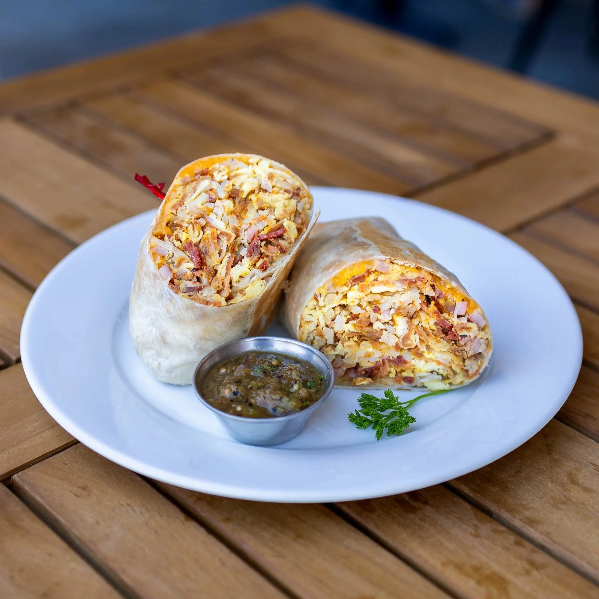 A burrito cut in half revealing scrambled eggs, bacon, and cheese on a white plate with a small dish of salsa and a sprig of cilantro, placed on a wooden table.