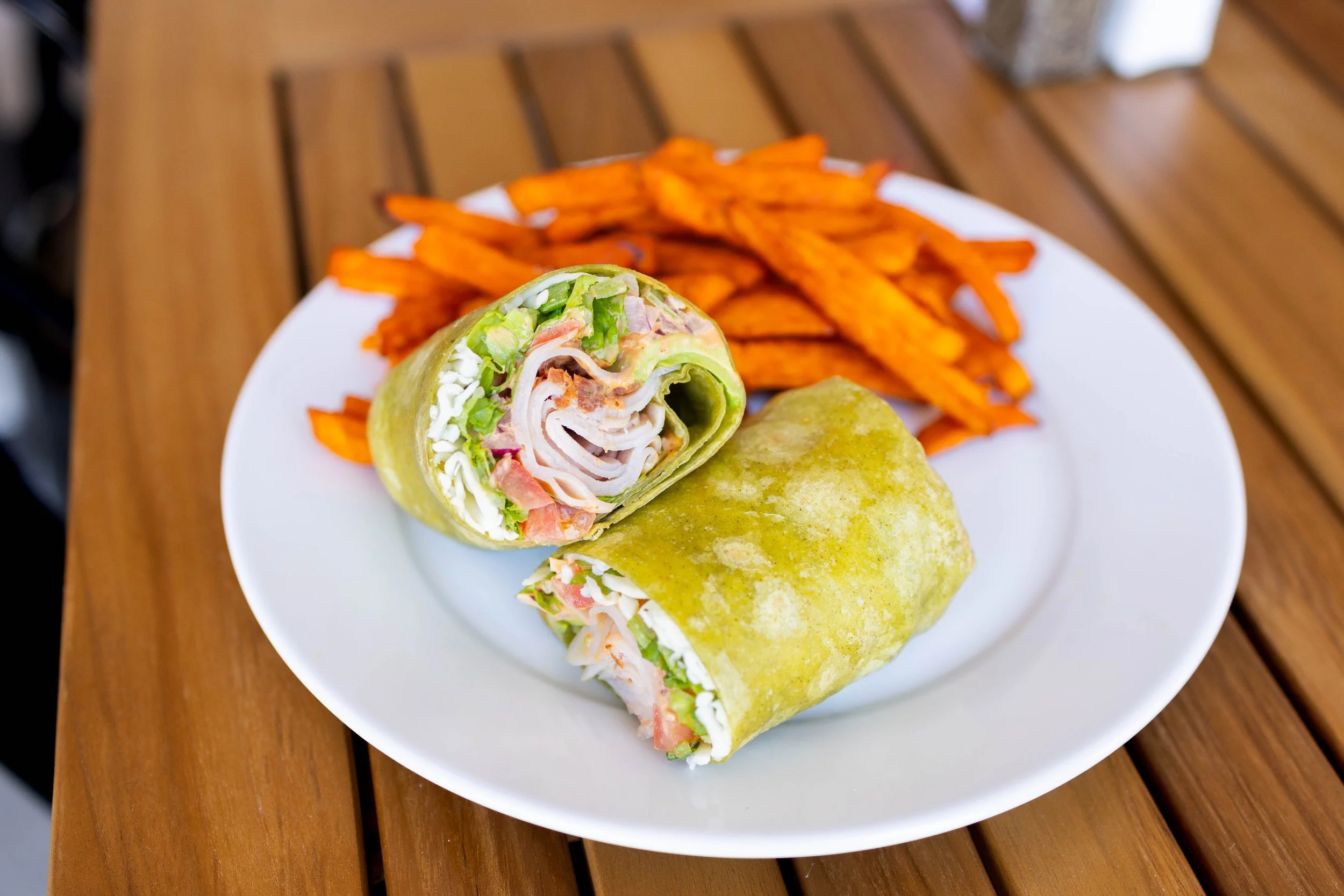 A plate of a veggie wrap cut in half, with orange sweet potato fries on the side, on a wooden table.