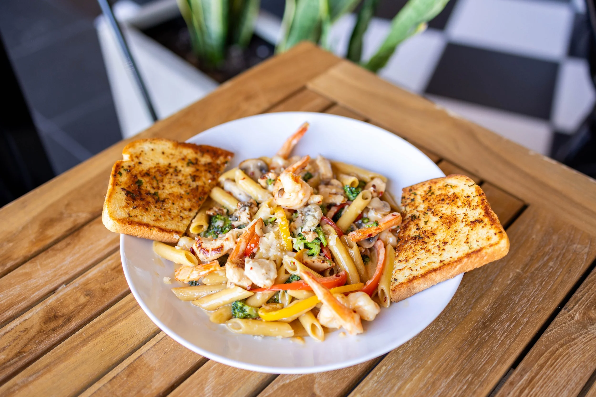 A white bowl of pasta with shrimp, vegetables, and cheese, served with two slices of toasted garlic bread on a wooden table.