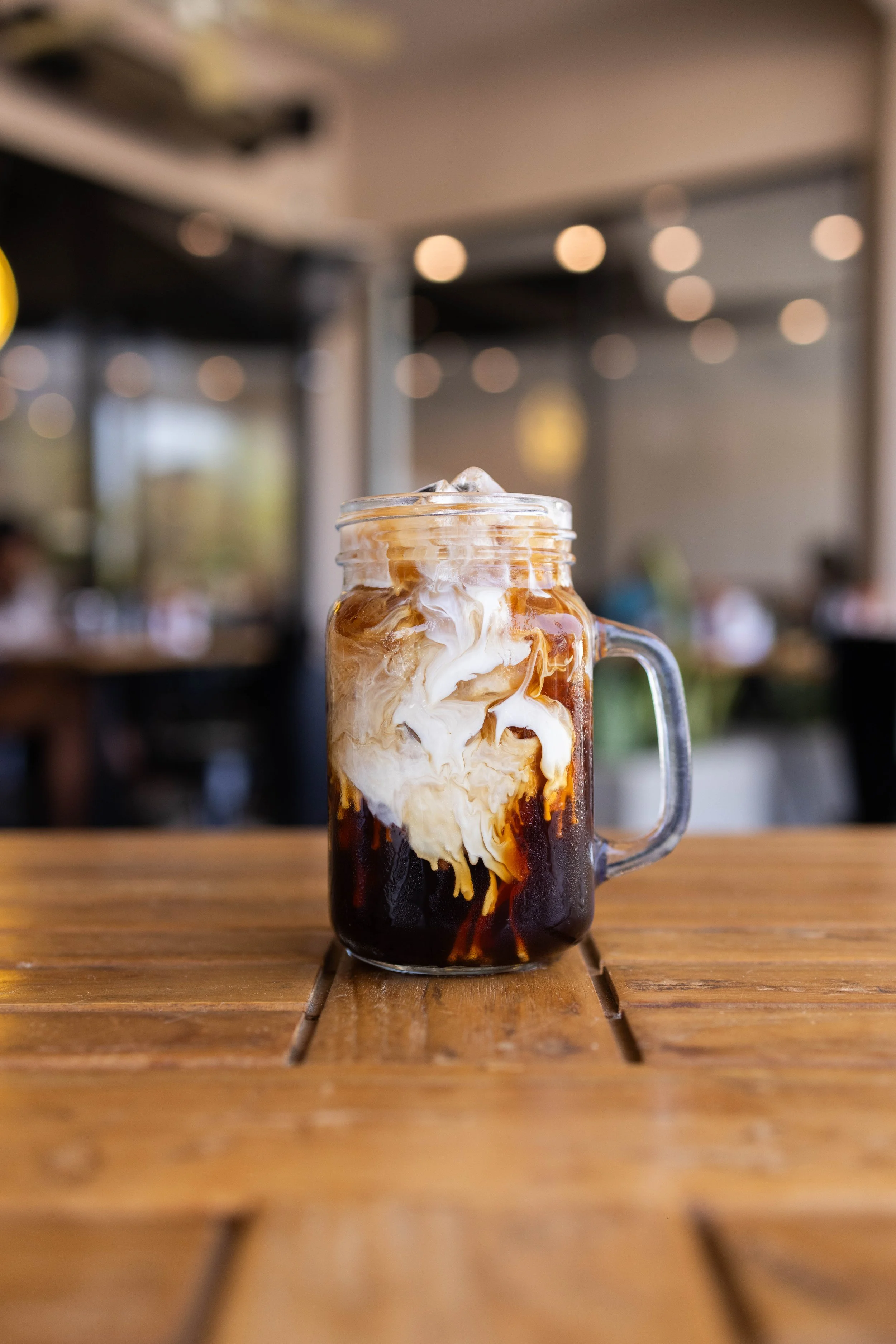 A glass jar mug filled with iced coffee and cream, with swirls of cream mixing into the dark coffee, placed on a wooden table in a cafe setting with blurred background.