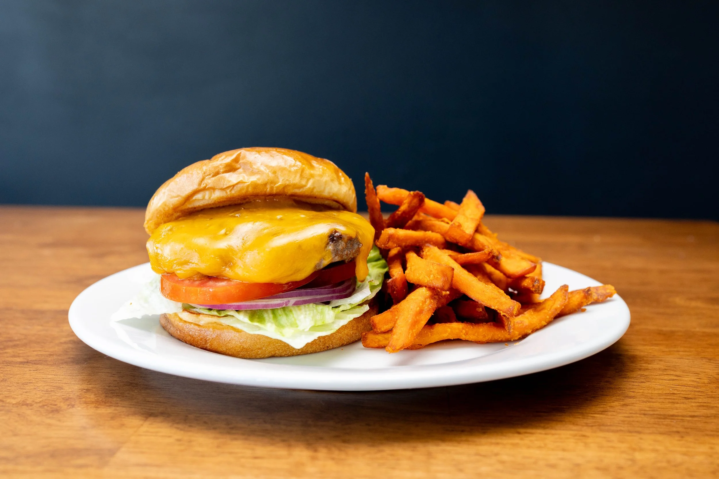 Cheeseburger with lettuce, tomato, onion, cheese, and a bun, served with sweet potato fries on a white plate.