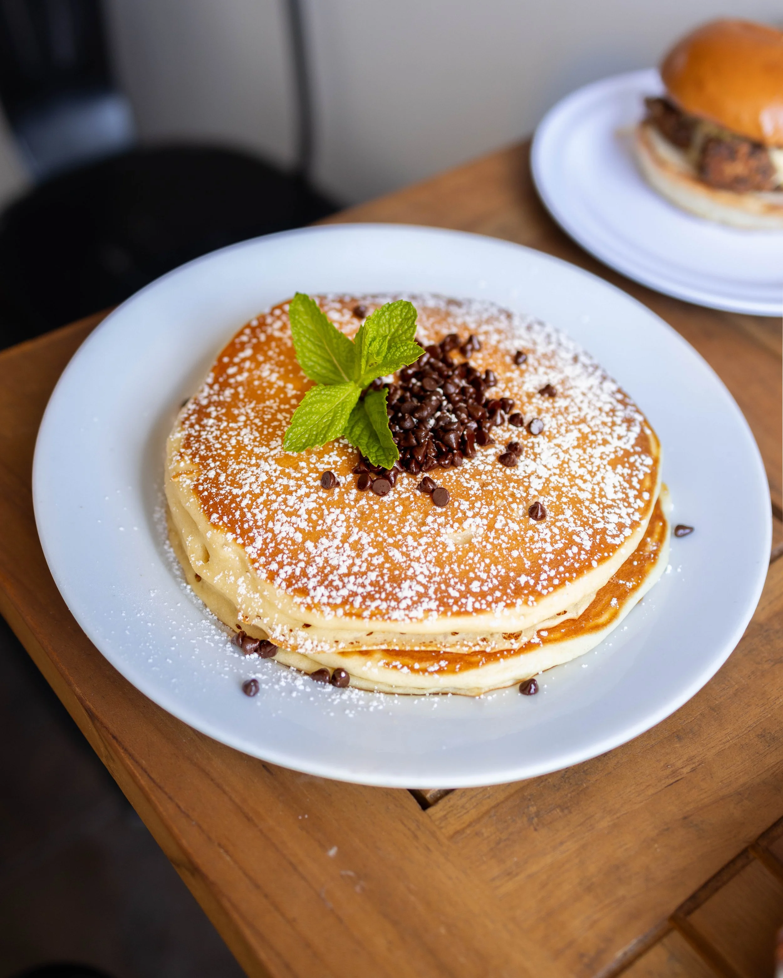 A plate of stacked pancakes topped with powdered sugar, chocolate chips, and a sprig of mint on a white dish on a wooden table.