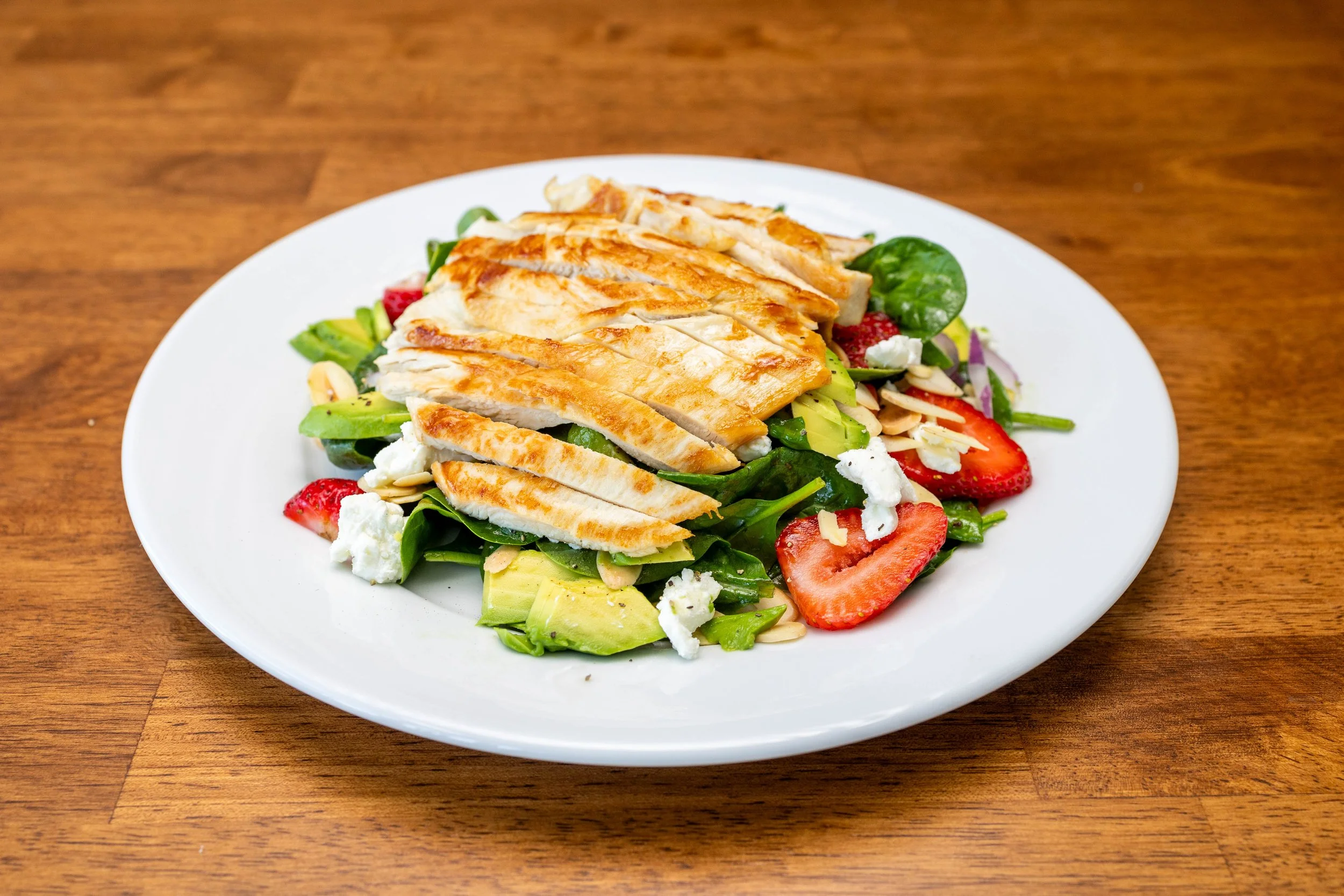 Grilled chicken salad with strawberries, spinach, avocado, feta cheese, and sliced almonds on a white plate on a wooden table.