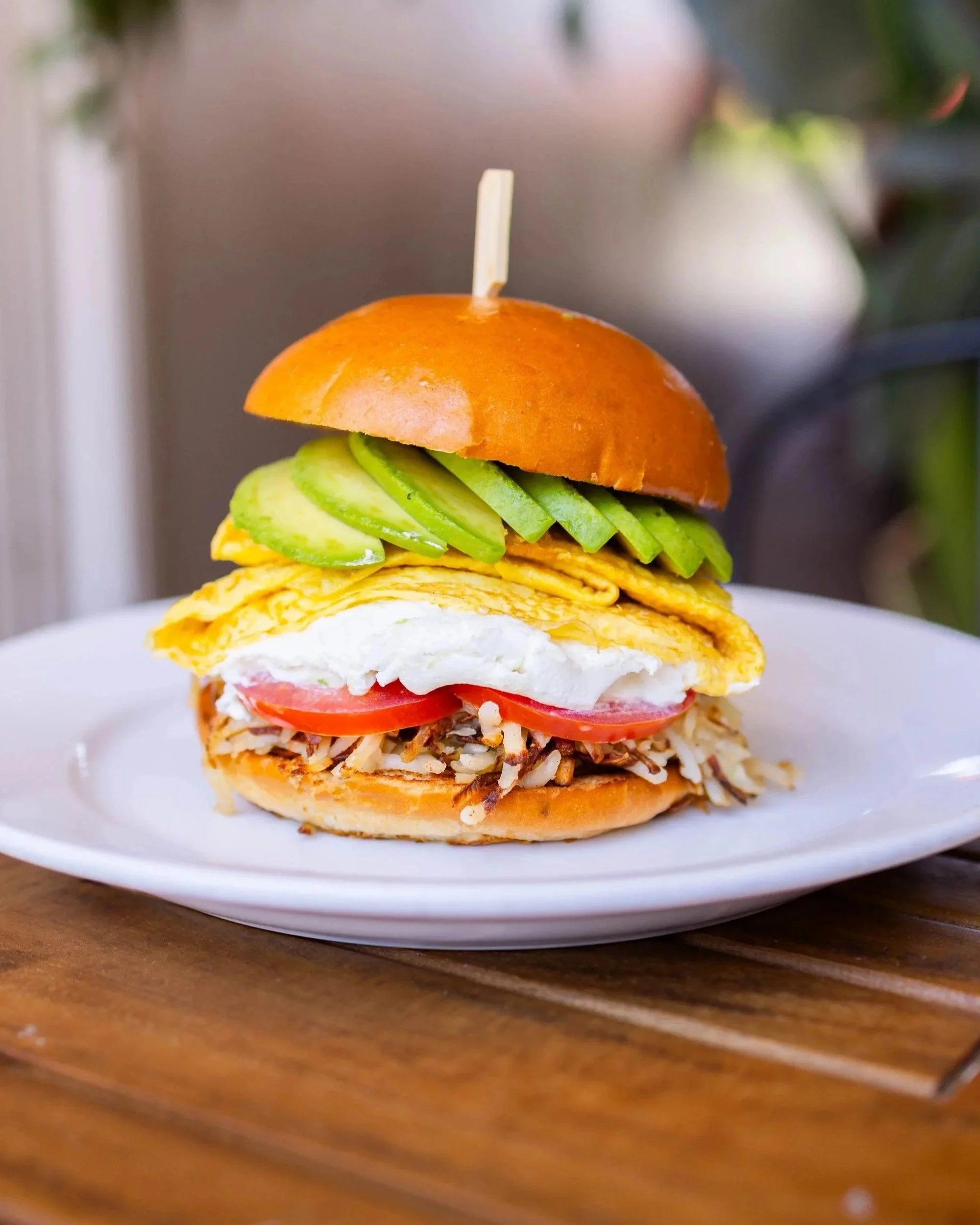 Stacked sandwich on a white plate with sliced avocado, scrambled eggs, tomatoes, shredded chicken, and a bun on top, with a skewer through it.